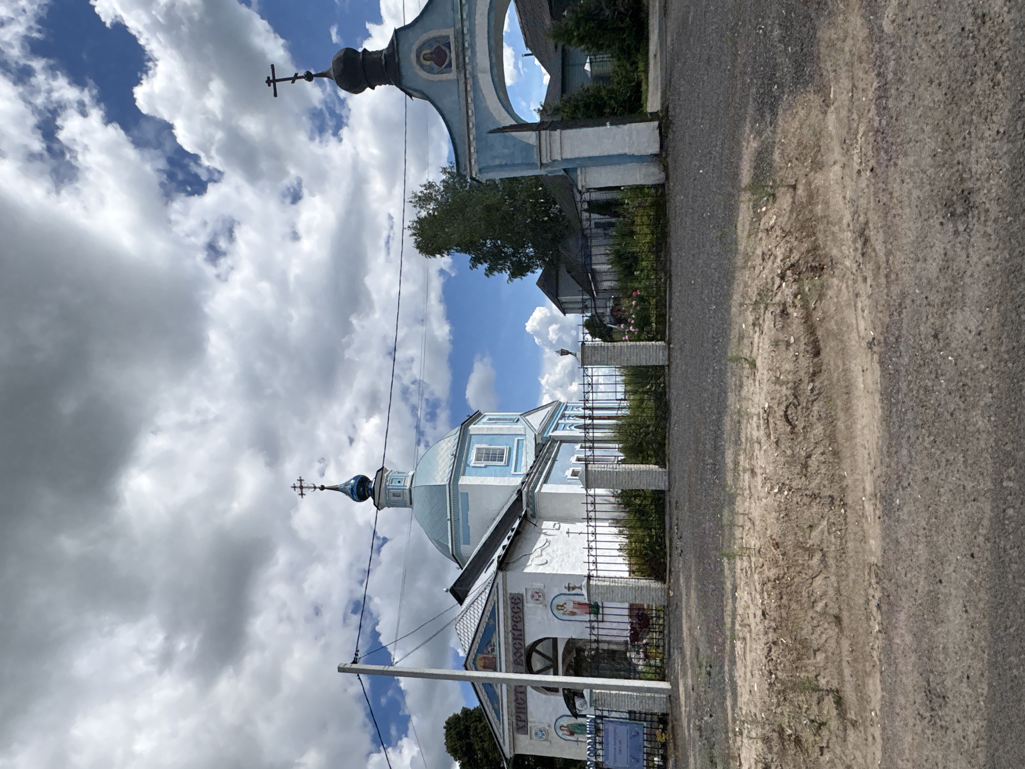 The image shows an Orthodox church with a blue dome and cross, located in the village of Глебовское, Russia. The church is white with blue trim and features several religious icons painted on its exterior walls. A fence with decorative pillars encloses the church grounds, where a small garden with flowers is visible. To the right of the church, a blue archway with a religious icon on top serves as an entrance. The church is situated on a gravel road, with a dirt embankment in the foreground. The sky is partly cloudy with a mix of blue sky and white clouds.