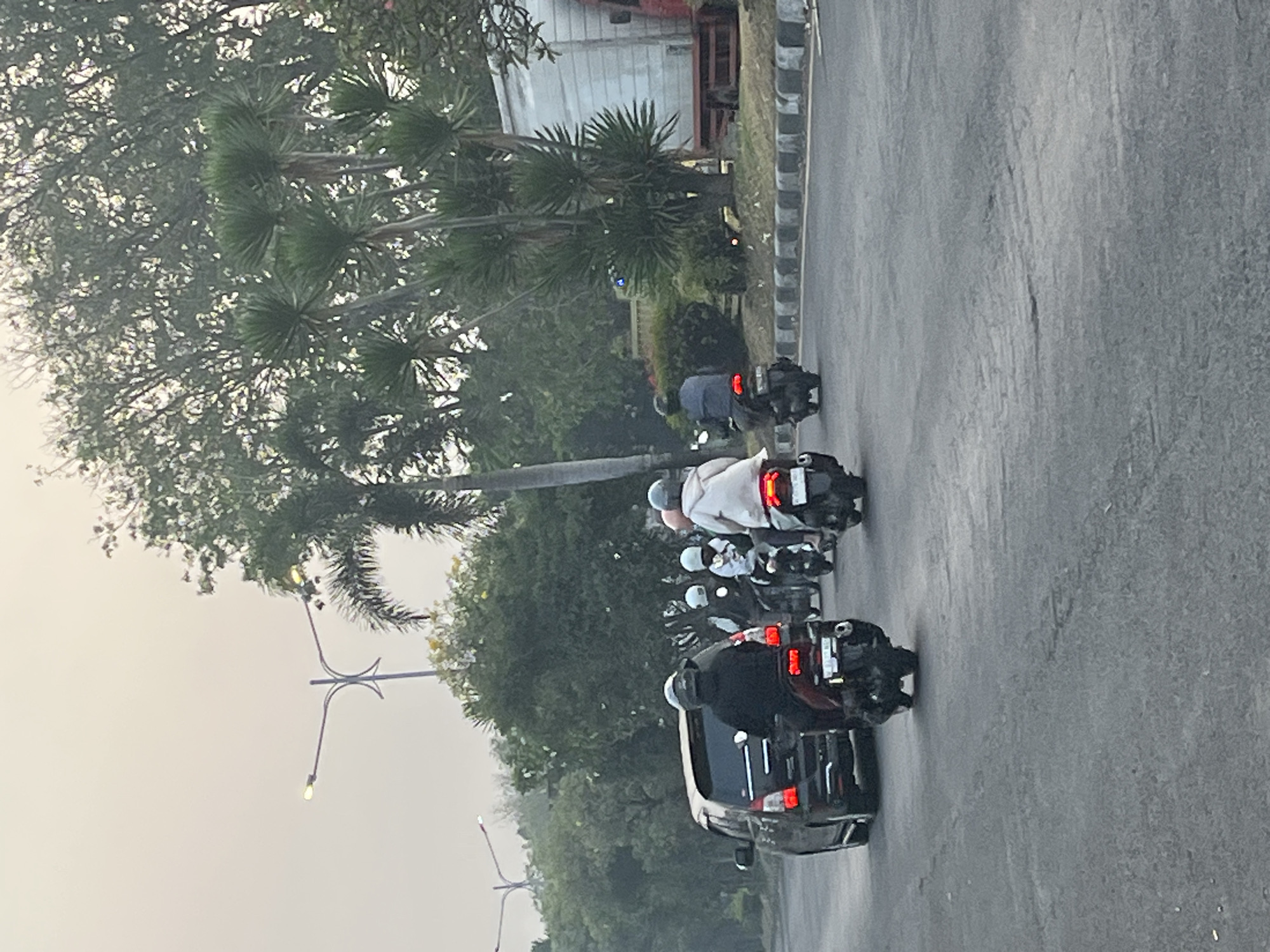 The image shows a road in Kludan, Indonesia, with several motorcycles and a car. The sky is overcast, and the trees are lush green. A group of people are riding motorcycles, most wearing helmets. One car is visible in the foreground. The scene appears to be a normal street scene in a tropical or subtropical climate.