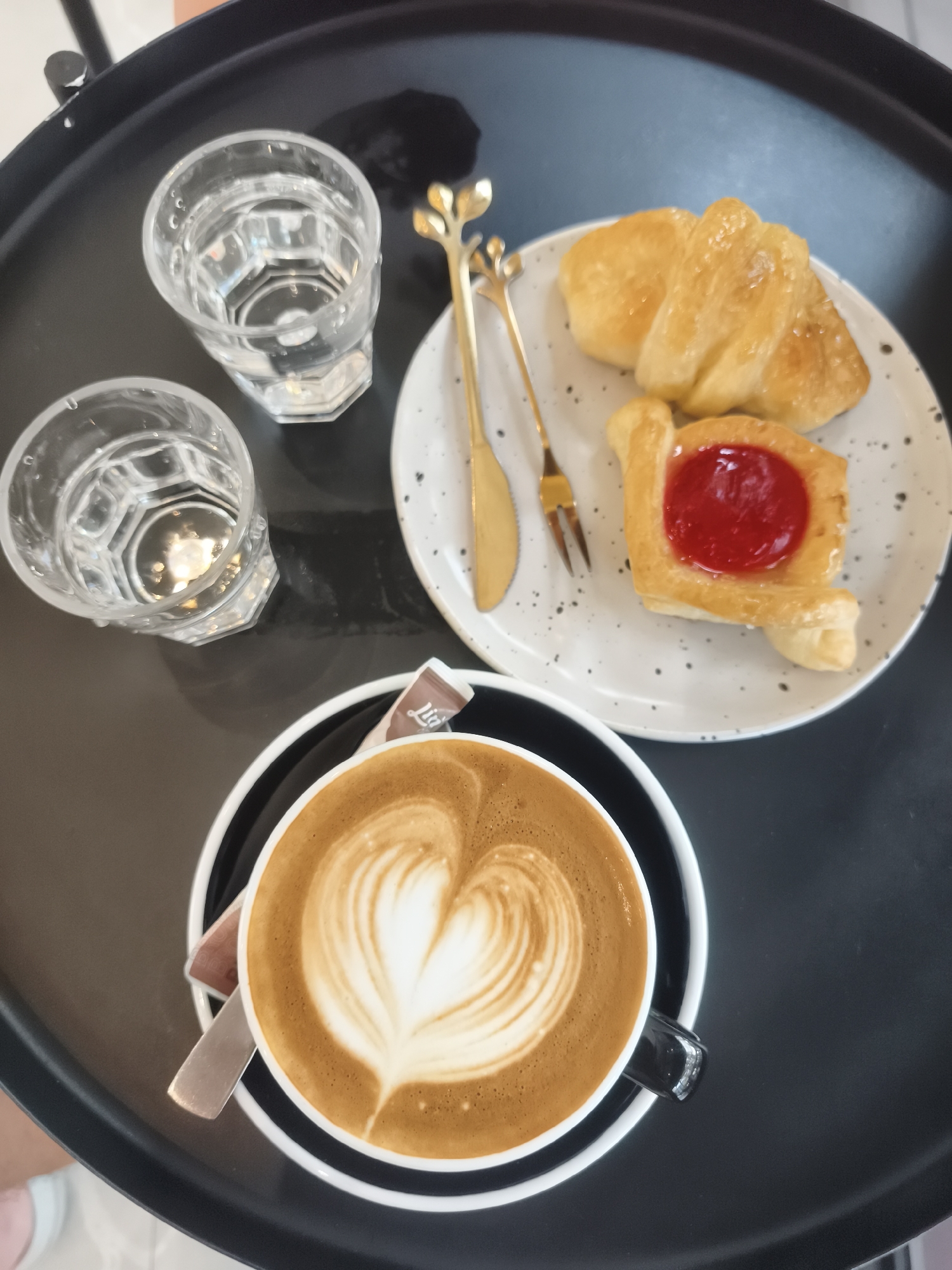A top-down, close-up shot captures a cafe scene on a black tray, likely in Ko Samui District, Thailand. A cappuccino with intricate latte art resembling a heart sits in a black mug on a saucer. Beside it, a small sachet of sugar, labeled "Lid," peeks out. To the right, a white plate holds two pastries: a golden croissant and a square pastry topped with red jam. Two glasses of water, one slightly closer to the viewer, are positioned on the left. Gold cutlery, a fork and a knife, rest on the plate. The tray sits on a surface, possibly a table, with a hint of a person's leg in the lower left corner wearing white shoes. The lighting suggests an indoor setting, possibly a cafe.