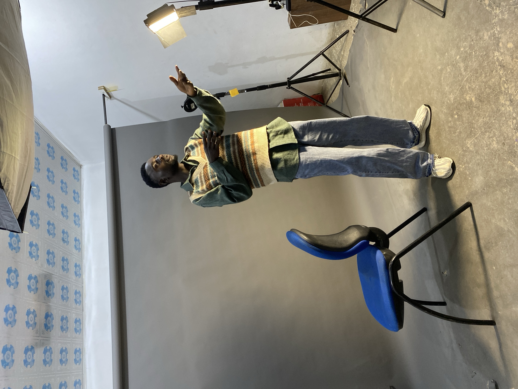 A man stands in a room with a blue chair and a gray backdrop. He is wearing jeans, a green jacket, and a striped sweater vest. The room has a patterned wall on the left side and a light stand in the background. The location is Jos, Nigeria. The man has his right hand raised.