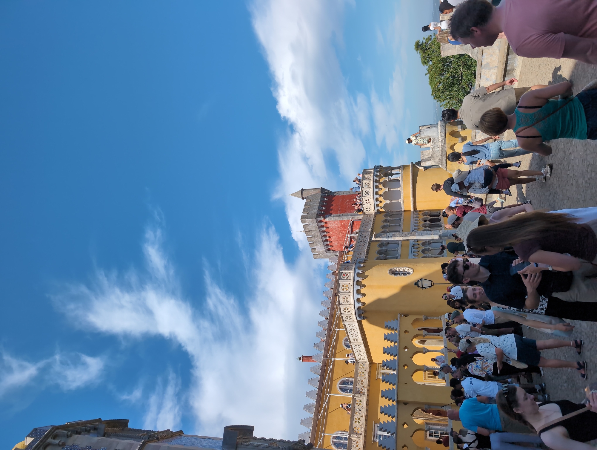 The image shows the Pena Palace in Sintra, Portugal. A group of tourists stands near the palace, some interacting with each other and their phones. The architecture of the palace is prominently displayed, with its yellow and red facade, ornate details, and towers. The sky is blue with some clouds.
