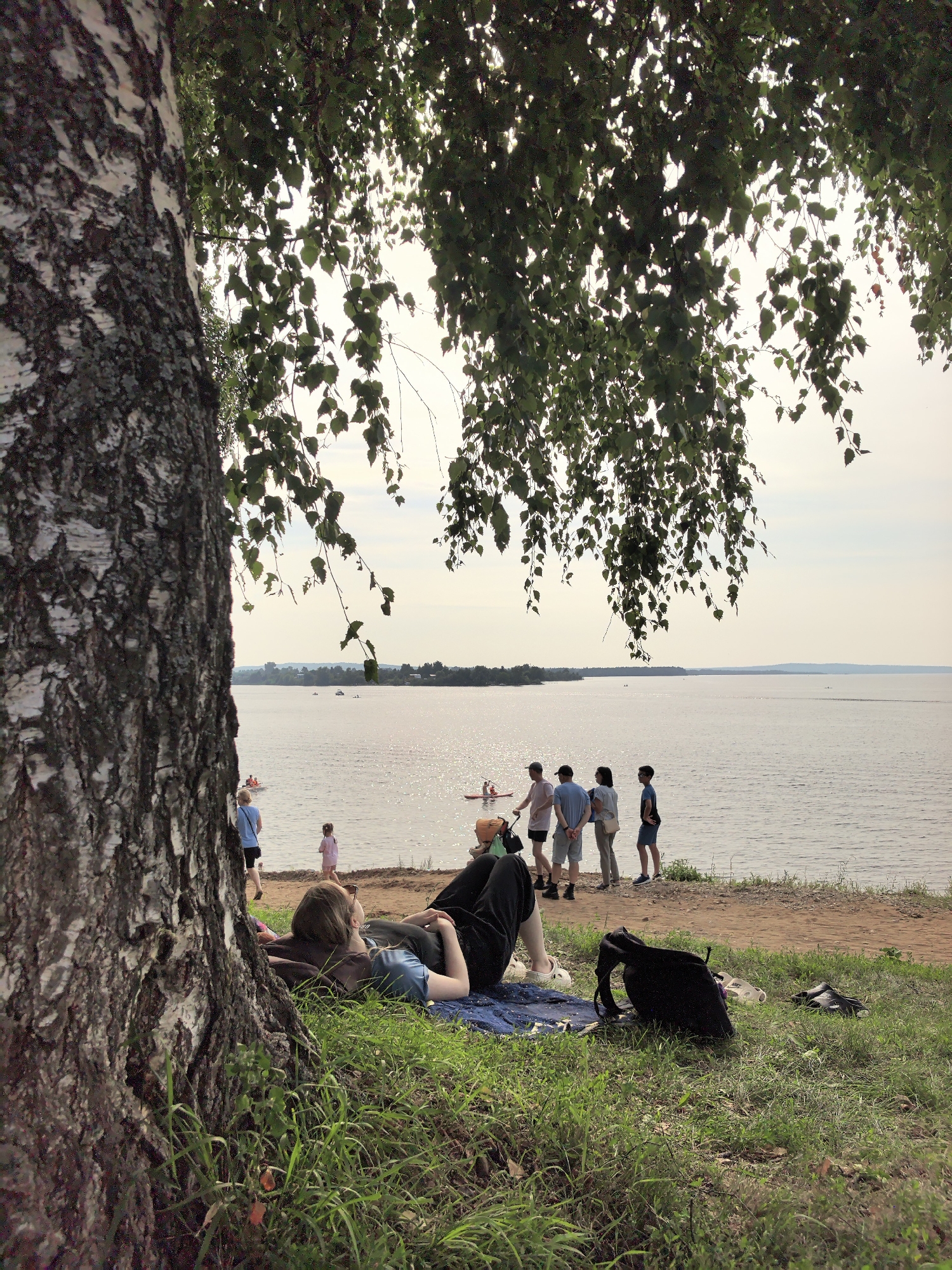 The image depicts a sunny day at a lakeside location in the city of Osa, Russia. In the foreground, a woman is lying on her back on a blue mat, her head resting on a dark cushion, under the dappled shade of a birch tree. A black backpack and a pair of shoes are placed on the grass beside her. The background shows a wide expanse of water, with several people standing near the shore, some on a sandy beach and others in the shallow water. Two individuals are on paddleboards in the water, and a small child in a pink dress is walking on the beach. Further in the distance, a treeline borders the horizon, with a hazy sky overhead. The overall atmosphere is one of leisure and outdoor recreation.