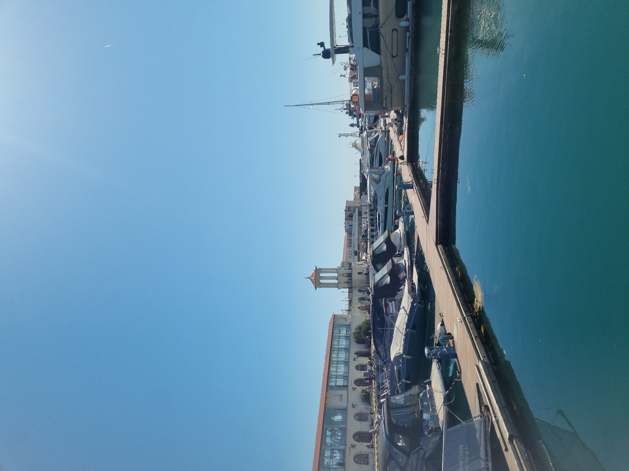 The image shows a marina in Sochi, Russia, under a clear blue sky. A row of docked yachts is visible alongside a concrete pier. The water is a turquoise color. Buildings with orange roofs and arched windows are situated adjacent to the marina, and a tower structure is visible further inland. The boats are covered in blue covers. A small plane is visible high in the sky.