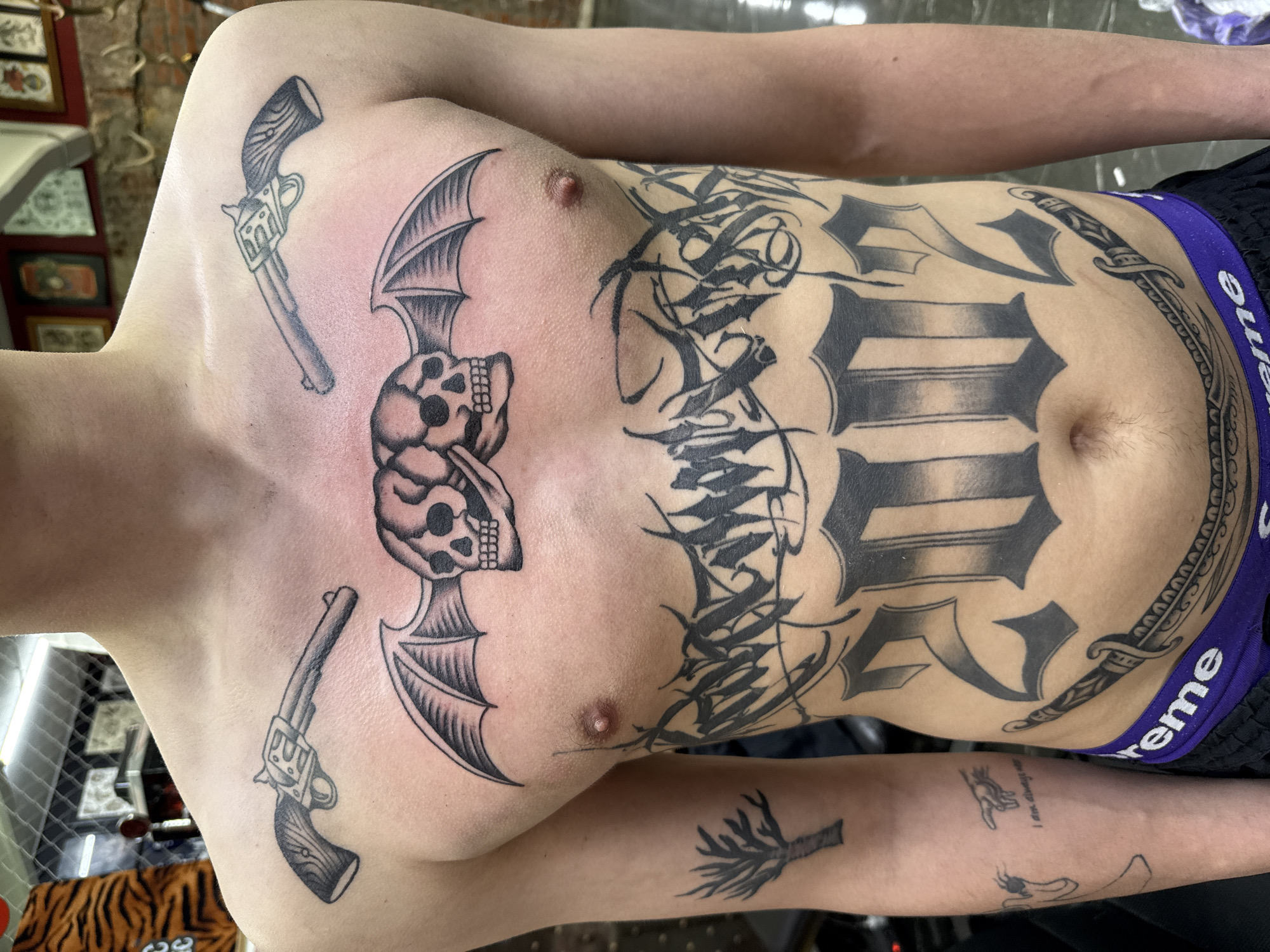 A person is photographed from the upper chest to the upper thighs. They have numerous black tattoos, including skulls with bat wings on the chest, guns on the upper chest, and various letters, numbers, and other symbols on the abdomen. They are wearing black shorts with a purple waistband that says "Supreme." The picture appears to be taken in a tattoo parlor. A portion of a chair with a tiger print is visible in the bottom left corner. The location of the photograph is Moscow, Russia.