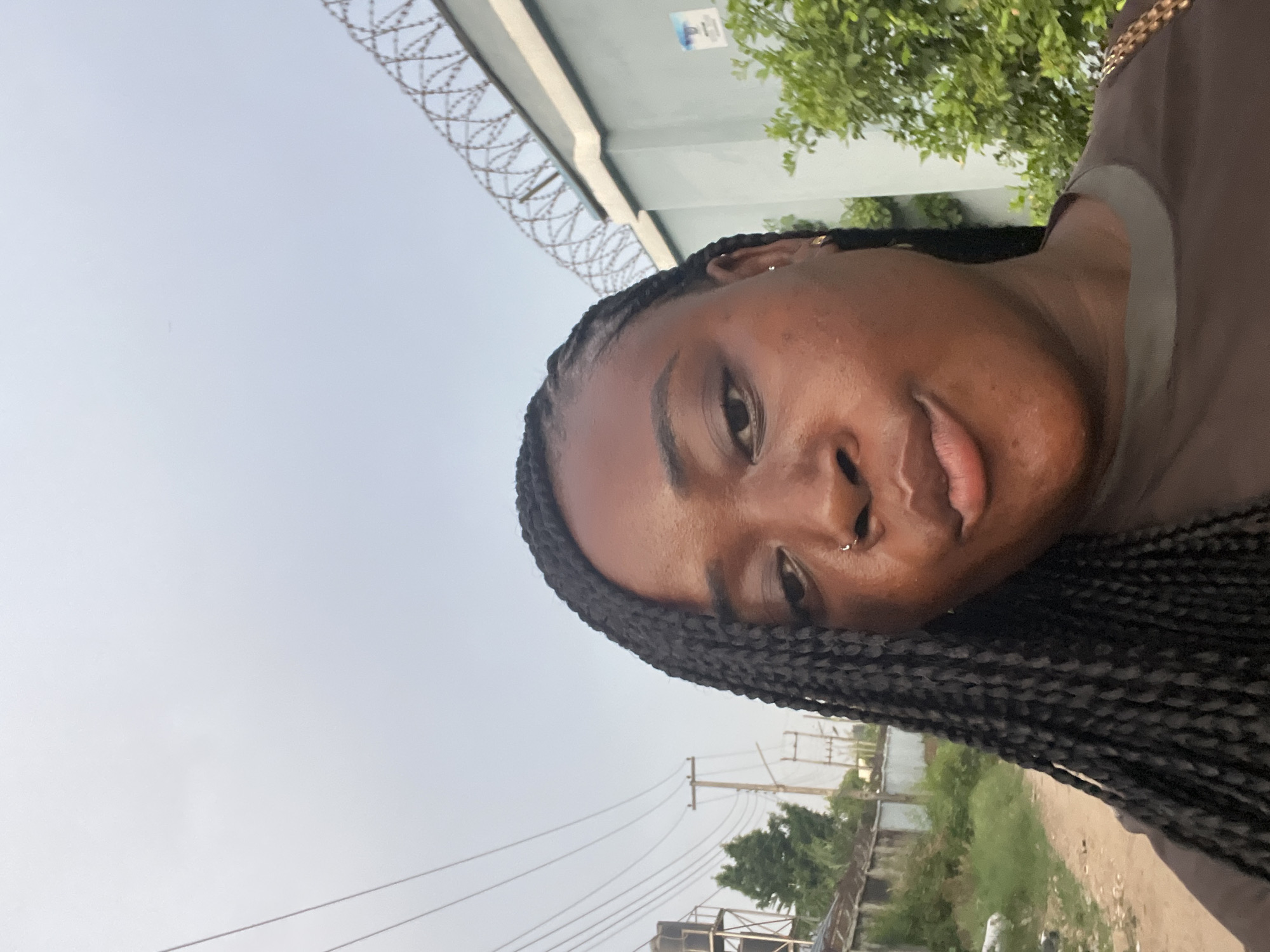 A medium close-up, angled shot shows a Black woman with dark braided hair facing the camera. She has a nose ring, and her expression is neutral. The background consists of a wall with barbed wire on top, trees, power lines, and what seems to be a cityscape. The location is possibly in Abuja, Nigeria, as specified.