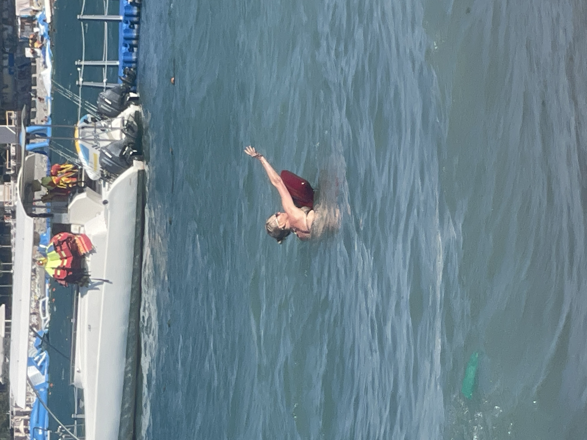 A person in a bikini is in the ocean, with a red floatation device beneath them. The person is wearing sunglasses and has their right arm extended upwards. In the background, a white boat with two outboard motors is docked at a blue platform. The water is choppy and a green object is visible floating in the foreground. The scene is likely Gelendzhik, Russia.