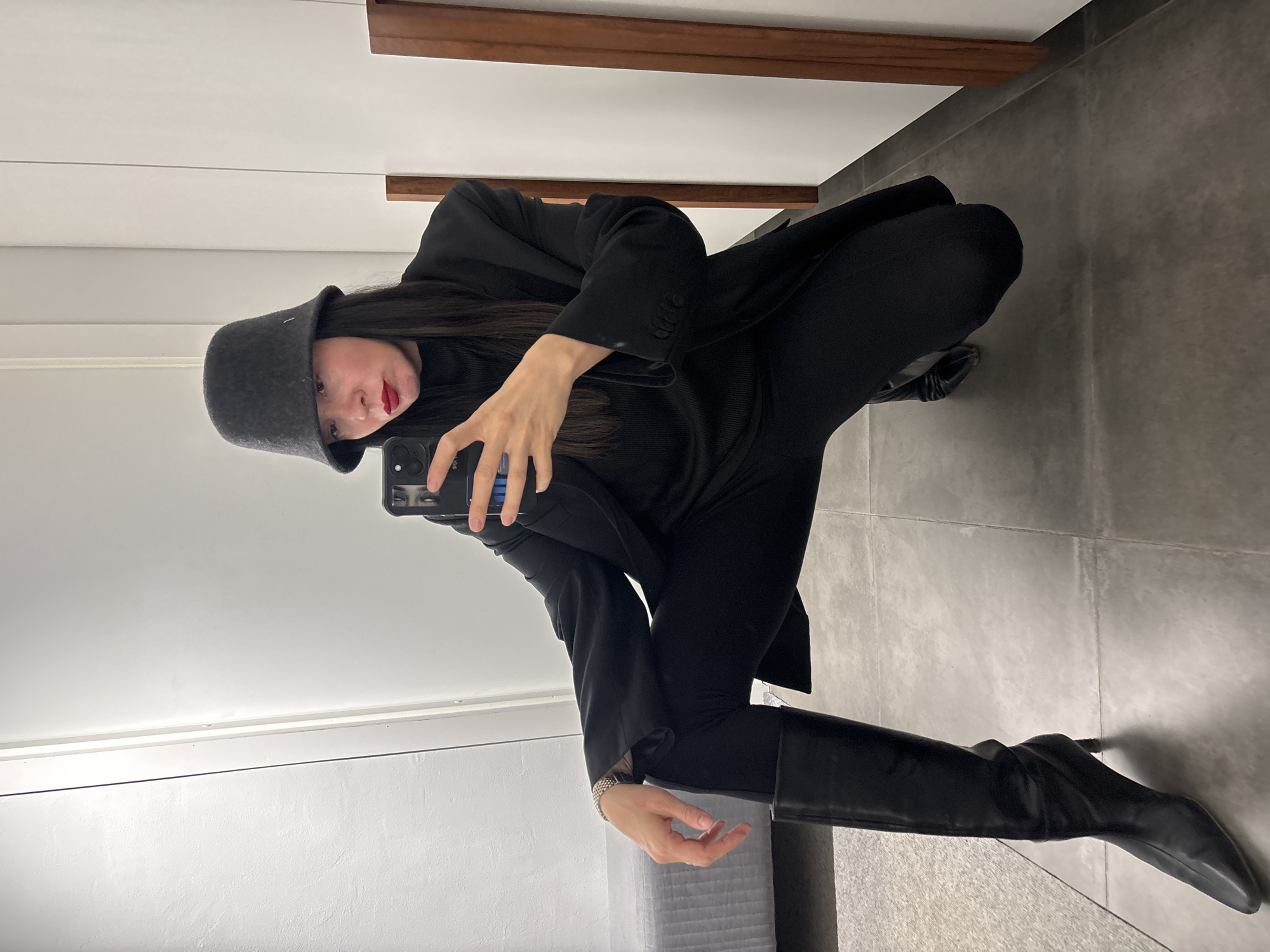 A woman is posing for a selfie in what appears to be a store fitting room. She is crouched down, wearing a black outfit consisting of a bucket hat, turtleneck sweater, blazer, pants, and high-heeled knee-high boots. She is holding her smartphone up with one hand and looking at the screen. Her lips are red. The walls of the fitting room are white and light grey. A dark wooden shelf is on the wall above her. The floor is grey tile. The location is presumed to be a store in Chaikovsky, Russia.