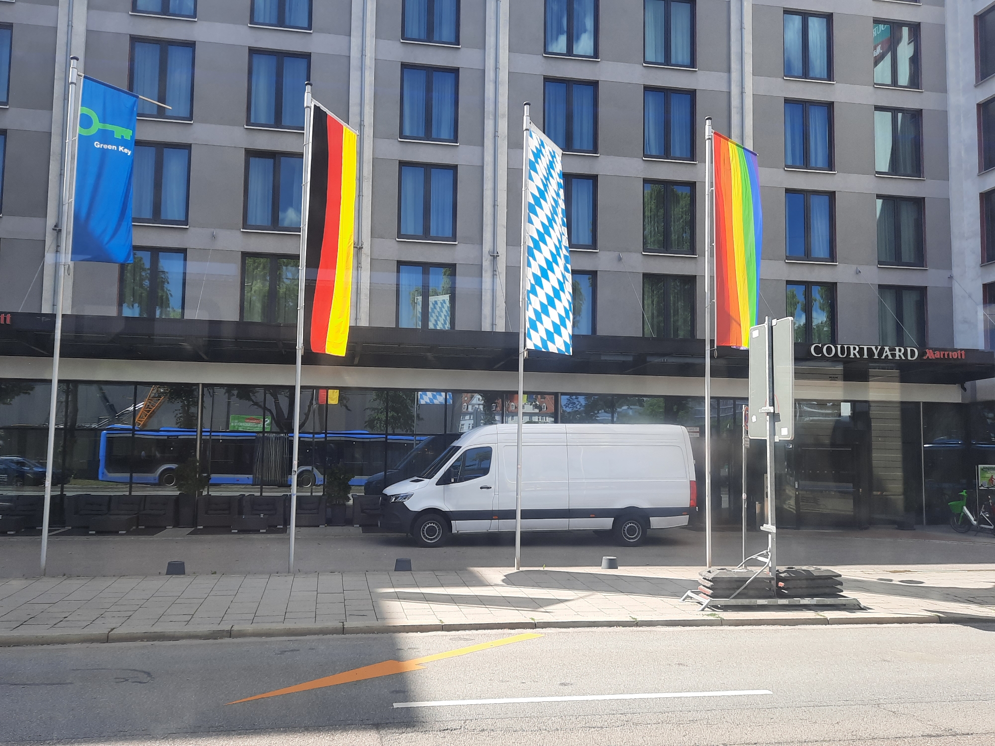 The image shows the exterior of a Courtyard Marriott hotel. In front of the hotel, there are four flags displayed on poles. From left to right, the flags are a blue flag with a green key symbol and the words "Green Key," the German flag (black, red, and yellow), the Bavarian flag (white and blue diamonds), and a rainbow flag. A white cargo van is parked on the sidewalk in front of the hotel. The hotel has a modern facade with many windows. In the reflection of the windows, a blue bus and a green bicycle are visible. The street in front of the hotel is paved with asphalt and has painted lane markings. There are several dark grey concrete barriers and stacks of black weights on the sidewalk near the flagpoles. The overall scene depicts an urban environment.