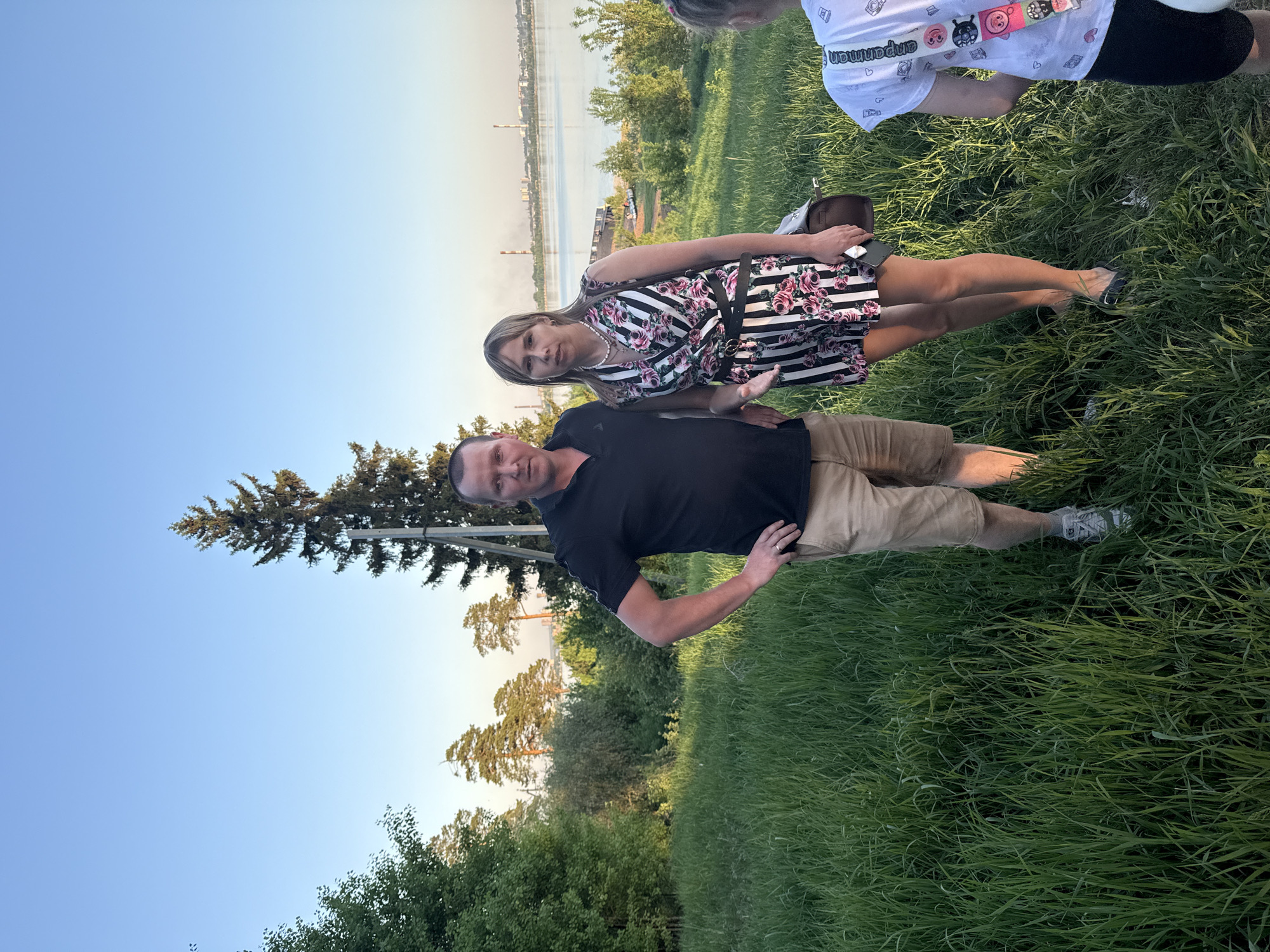 A man and a woman are standing in tall grass in an outdoor setting. The woman is wearing a striped and floral dress with a black belt. The man is wearing a black t-shirt and khaki shorts. There is a body of water in the background, and some buildings can be seen on the opposite shore. The environment appears to be a park or green space. The city in the background is Izhevsk, Russia.