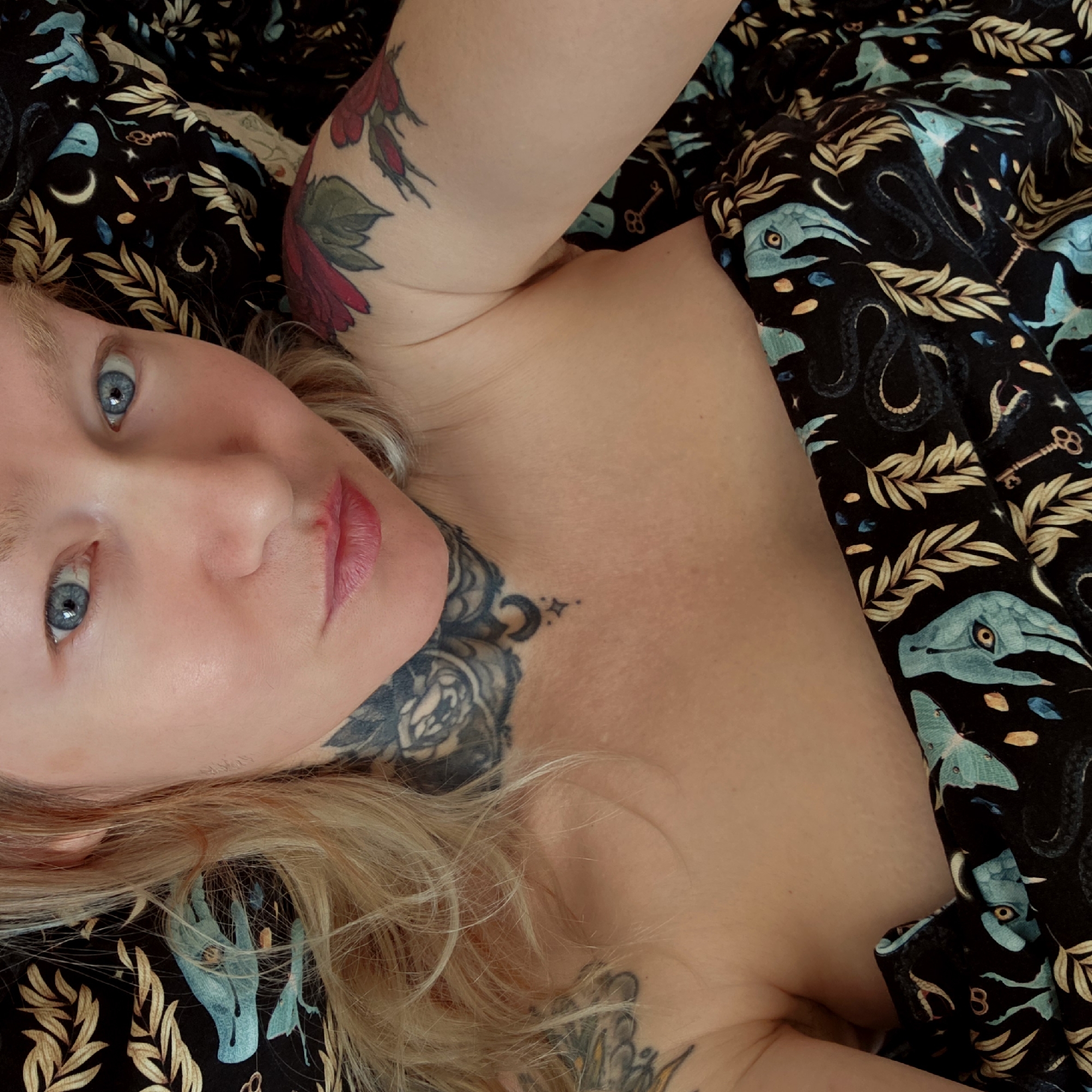 A fair-skinned woman with blue eyes and blonde hair is lying down. She has visible tattoos on her upper left arm and on her neck. She appears to be partially covered by a black fabric featuring a repeating pattern of blue and gold designs, including botanical elements, crescent moons, snakes, keys, and stylized animals. The scene does not contain any information suggesting its location in Saint Petersburg, Russia, or any other city.