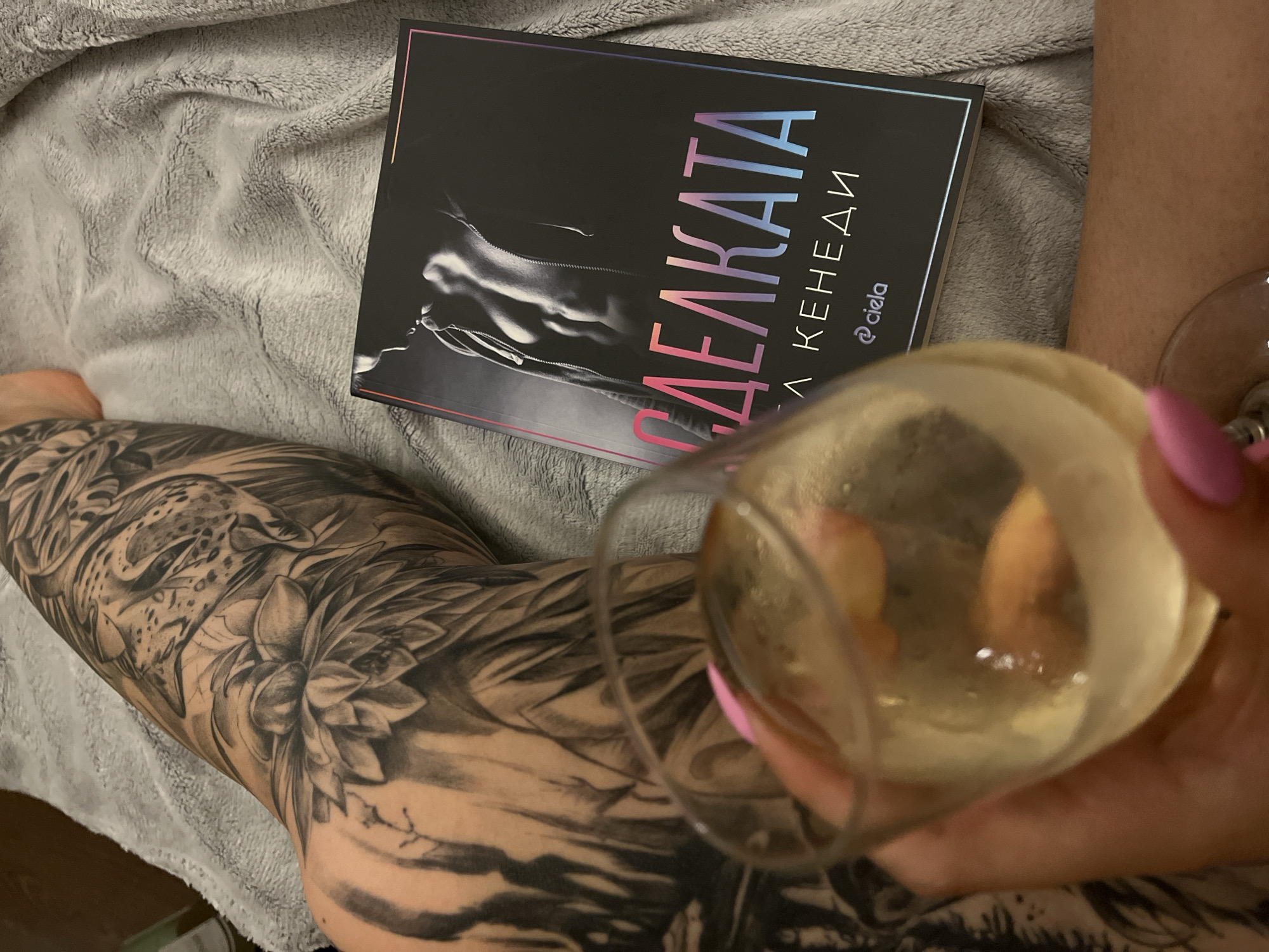 The image presents a close-up, top-down perspective of a person relaxing indoors in Veliko Tarnovo, Bulgaria. The scene features a person's leg adorned with a detailed black and gray tattoo, a book, and a hand holding a glass.
The leg is extensively tattooed with a large-scale design, prominently featuring the head of a leopard or jaguar, alongside intricate floral elements resembling tropical flowers and dense foliage. The tattoo's style is realistic and highly detailed, covering a significant portion of the leg.
Resting on a soft, light gray textured blanket is a book with a dark cover. The cover design includes a partial, silhouetted image of a person's back, possibly wearing a hooded garment. The title is visible in Cyrillic: "ДЕЛЕКАТА" (translating to "THE DELICATE" or "THE DELICATE ONE"), rendered in a striking pink-to-blue gradient. Below the title, the author's name "ЕЛ КЕНЕДИ" (ELLE KENNEDY) is also visible, along with the "ciela" publisher logo.
In the foreground, a hand with pink manicured nails holds a stemmed glass containing a pale yellow liquid, suggesting champagne or white wine. The glass is slightly out of focus, drawing attention to the book and tattoo. The overall setting conveys a relaxed and comfortable ambiance, indicative of a leisurely activity like reading and enjoying a beverage.