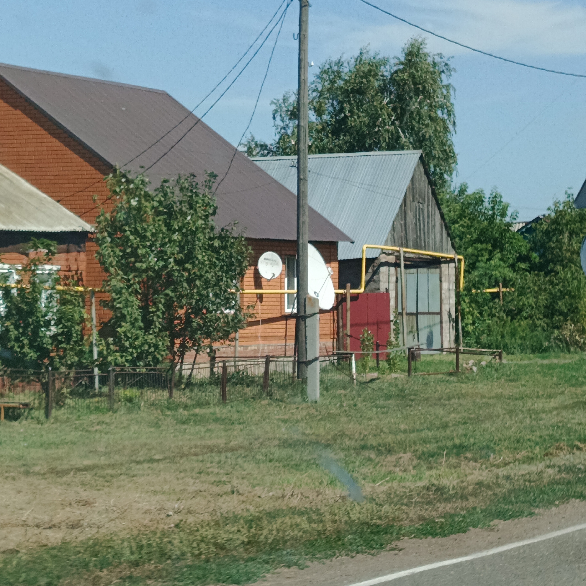 The image captures a rural scene with two houses and a grassy area in front of them. The house on the left is partially visible, constructed with red brick and a brown metal roof. Adjacent to it is a second house, primarily wooden with a corrugated metal roof, featuring a red-painted door and a light blue framed window. A yellow gas pipe is visible running along the exterior of this house. Two satellite dishes are mounted on the wooden house. A wooden utility pole with electrical wires stands in the foreground, slightly off-center. The area in front of the houses is a lawn with some scattered vegetation and a metal fence. The background consists of trees and a clear blue sky. The location is identified as Raznomoyka, Russia. There are no people or animals visible in the image.