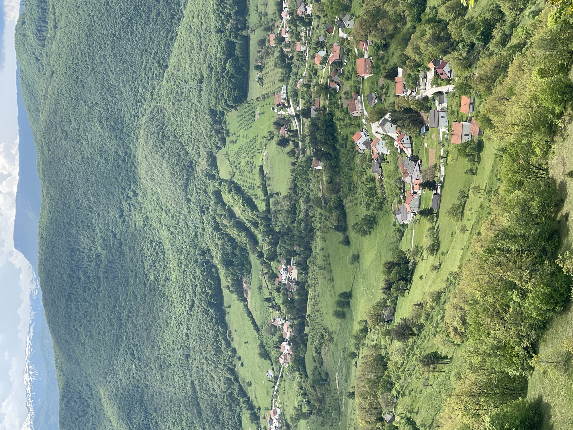 The image shows a landscape view in Gmići, Bosnia and Herzegovina. The left side is dominated by a densely forested mountainside. To the right of the mountainside is a valley with green fields and sparsely distributed trees. Within this valley, several clusters of buildings, likely houses, are visible, forming small settlements. The roofs of the houses appear mostly red or gray. A winding road or path connects the settlements. The overall impression is that of a rural, mountainous area with dispersed residential areas.