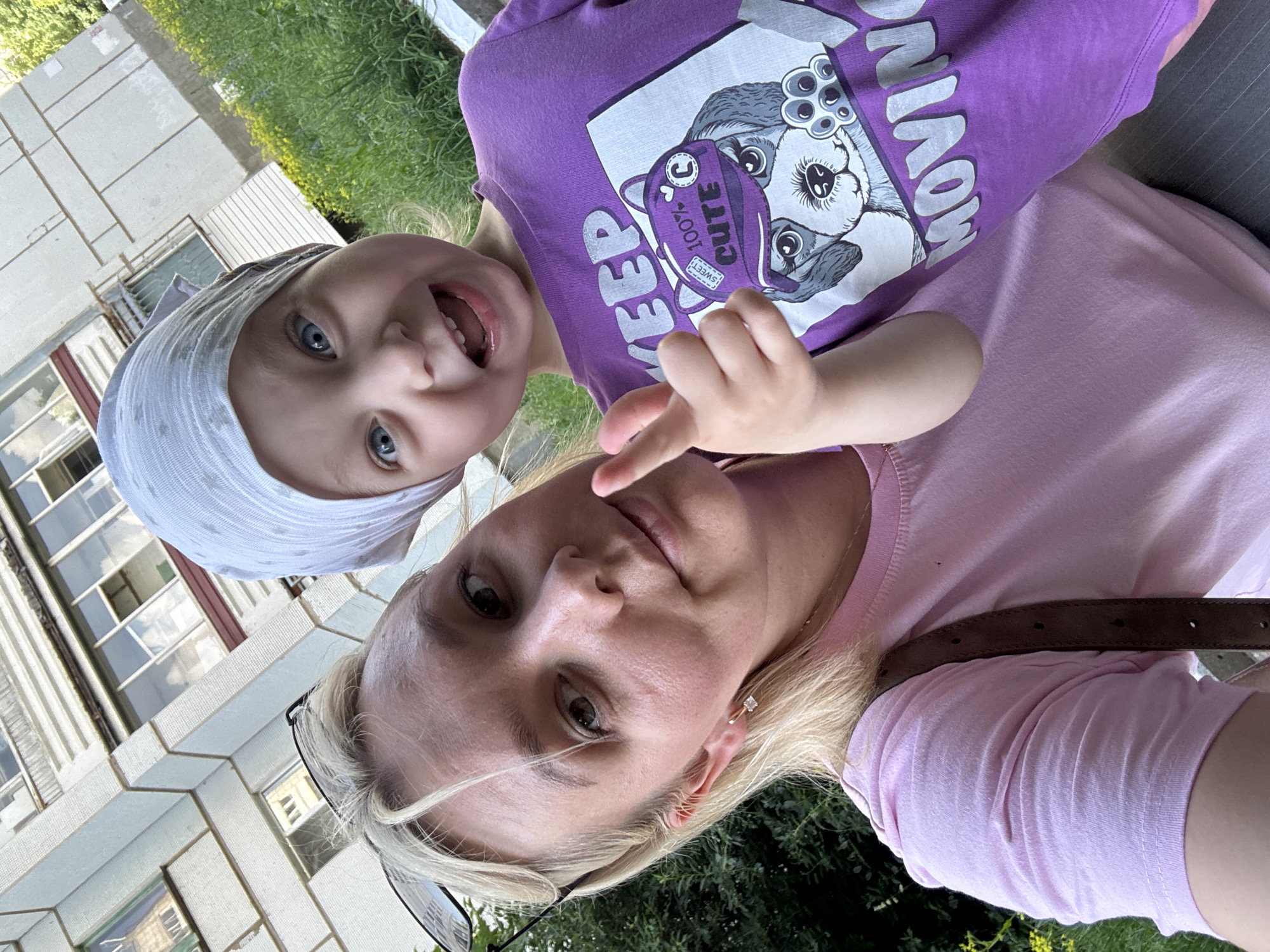 The image is a selfie taken outdoors, possibly during daytime given the bright lighting. A woman with blonde hair and glasses is in the foreground, wearing a pink t-shirt and a brown belt. Next to her, a toddler with blue eyes and a white head covering is looking at the camera with a wide smile. The toddler is wearing a purple t-shirt with a graphic print of a dog.
The background shows greenery and parts of a building with many windows, suggesting an urban or suburban setting. The people appear to be in good spirits, with the toddler exhibiting an excited expression. The text on the toddler's shirt is partially visible, reading "KEEP 100% CUTE" and "SWEET". The overall scene conveys a casual and happy moment between a mother and her child.