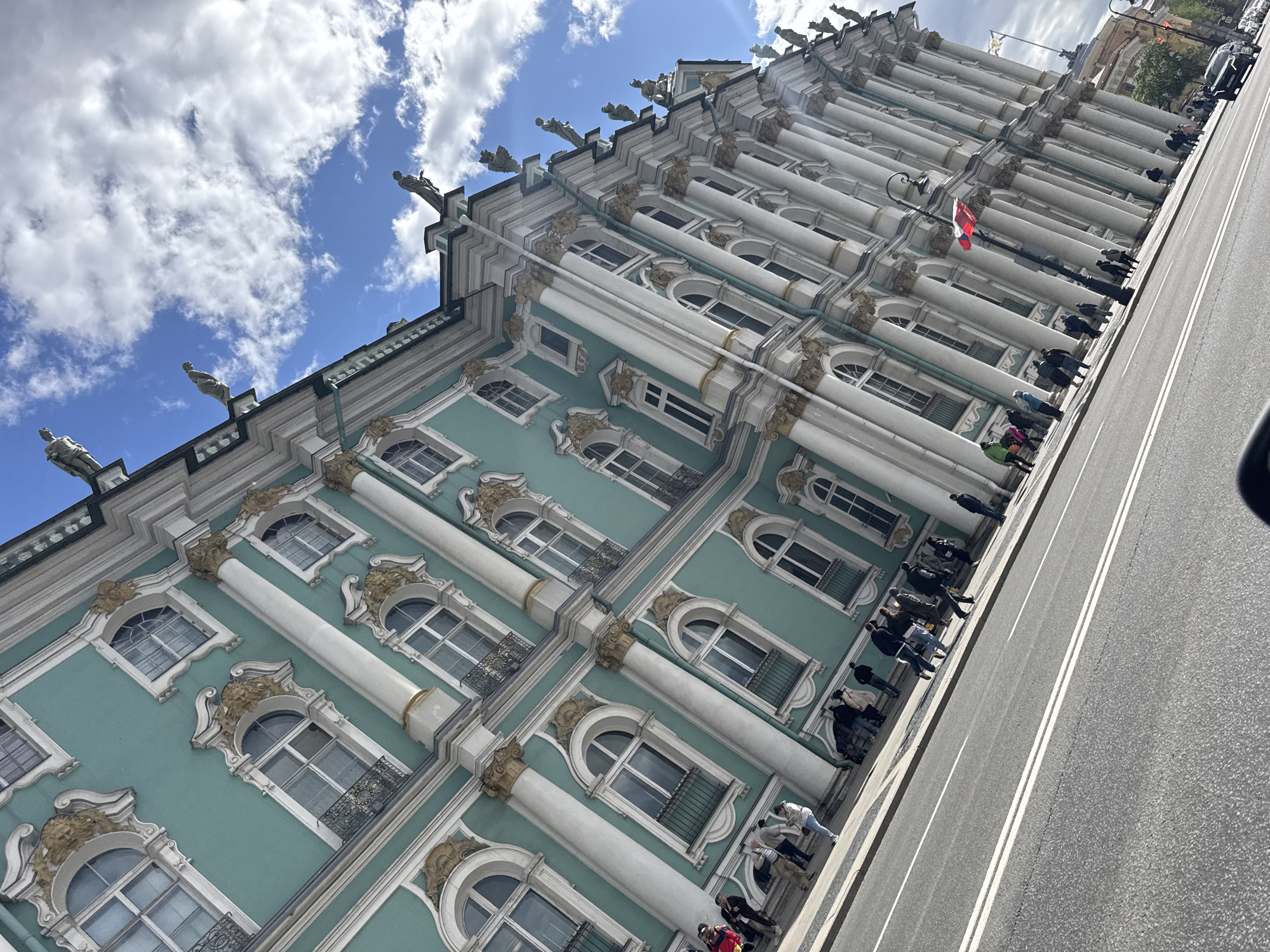 The image shows a street-level view of the Winter Palace in Saint Petersburg, Russia. The pale blue and white facade of the building, with its ornate window details and classical columns, is prominent against a backdrop of a partly cloudy blue sky. A row of people stands along the sidewalk, possibly waiting to enter the building, which is a branch of the Hermitage Museum. A road runs alongside the palace, with white lane markers visible. There is a flag pole with a red and white flag attached to the facade.