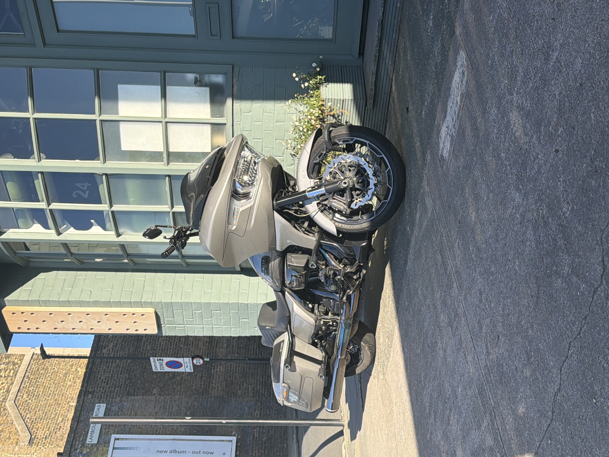A grey, modern touring motorcycle is parked on a paved surface in front of a building on a bright, sunny day in London, United Kingdom. The motorcycle, possibly a Harley-Davidson or similar, features a large front fairing, chrome engine components, a prominent exhaust, and saddlebags. The front wheel has intricate spokes, and "Brembo" is visible on the brake caliper, indicating high-performance brakes.
Behind the motorcycle is a building with a muted green facade and large windows with multiple panes. A matching green door is also visible. The number "24" is displayed on one of the window frames. Small white flowers grow near the building's base, between the motorcycle and the wall. To the left, another building made of light-colored brick can be seen.
The scene is well-lit by direct sunlight, casting strong shadows from the motorcycle and buildings, indicating either late morning or early afternoon on a clear day. On the street to the left, several signs are visible. A street sign clearly reads "LAMBOURN ROAD," confirming the specific London location. Another sign indicates "Controlled ZONE B" with specific hours: "10.00-11.00 & 14.00-15.00 Mon-Fri." A circular red sign shows "20," denoting a speed limit. Partially visible at the bottom left is a white poster with the text "new album - out now." No people are visible, and the motorcycle appears to be stationary.