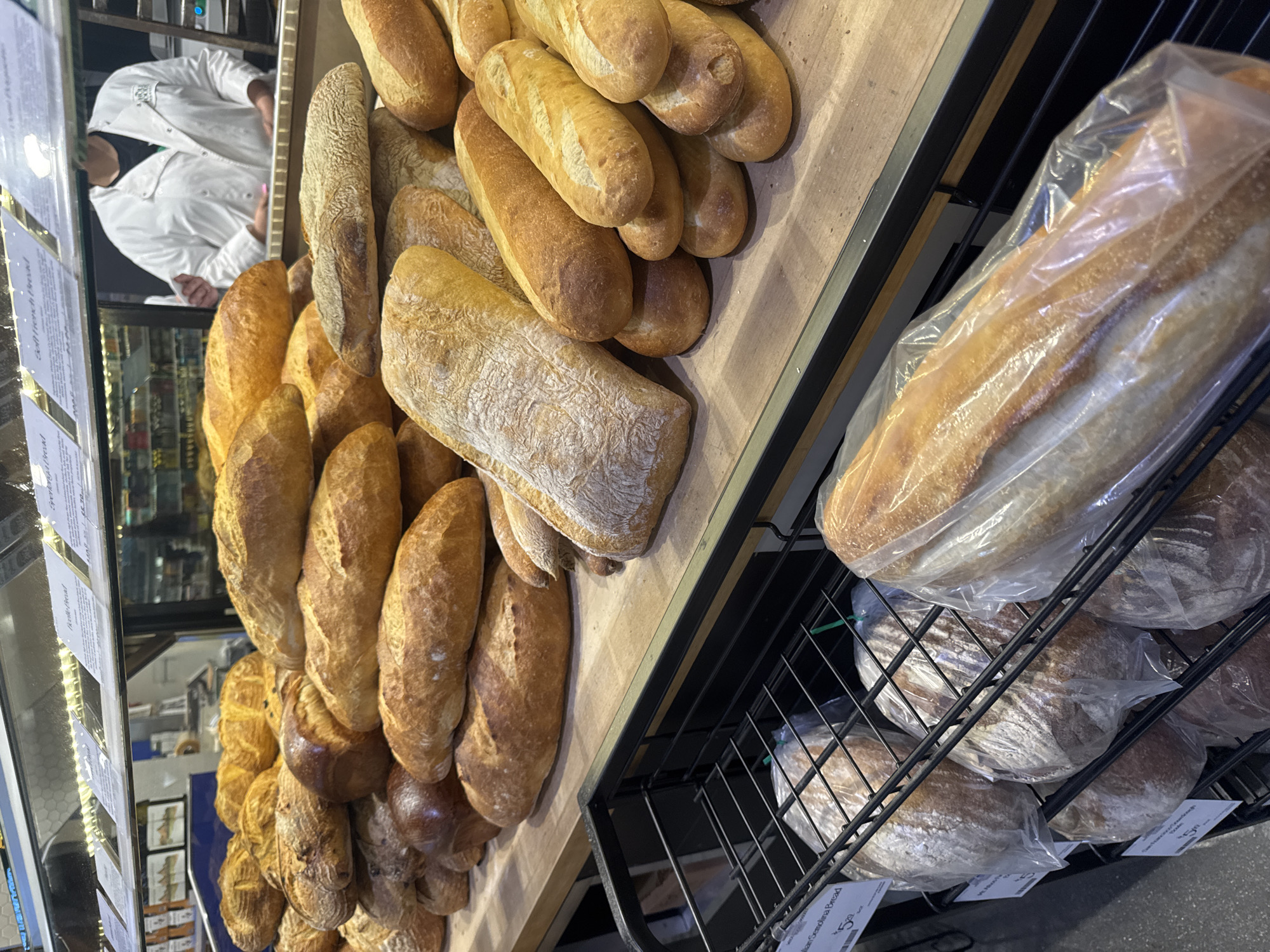 The image depicts a display of bread in a bakery or grocery store. A person wearing a white coat is visible behind a glass display. Assorted loaves of bread are arranged on shelves. Some loaves are individually packaged in clear plastic bags. Pricing tags are attached to the products on the shelves below. The location is assumed to be within Los Angeles, United States of America.