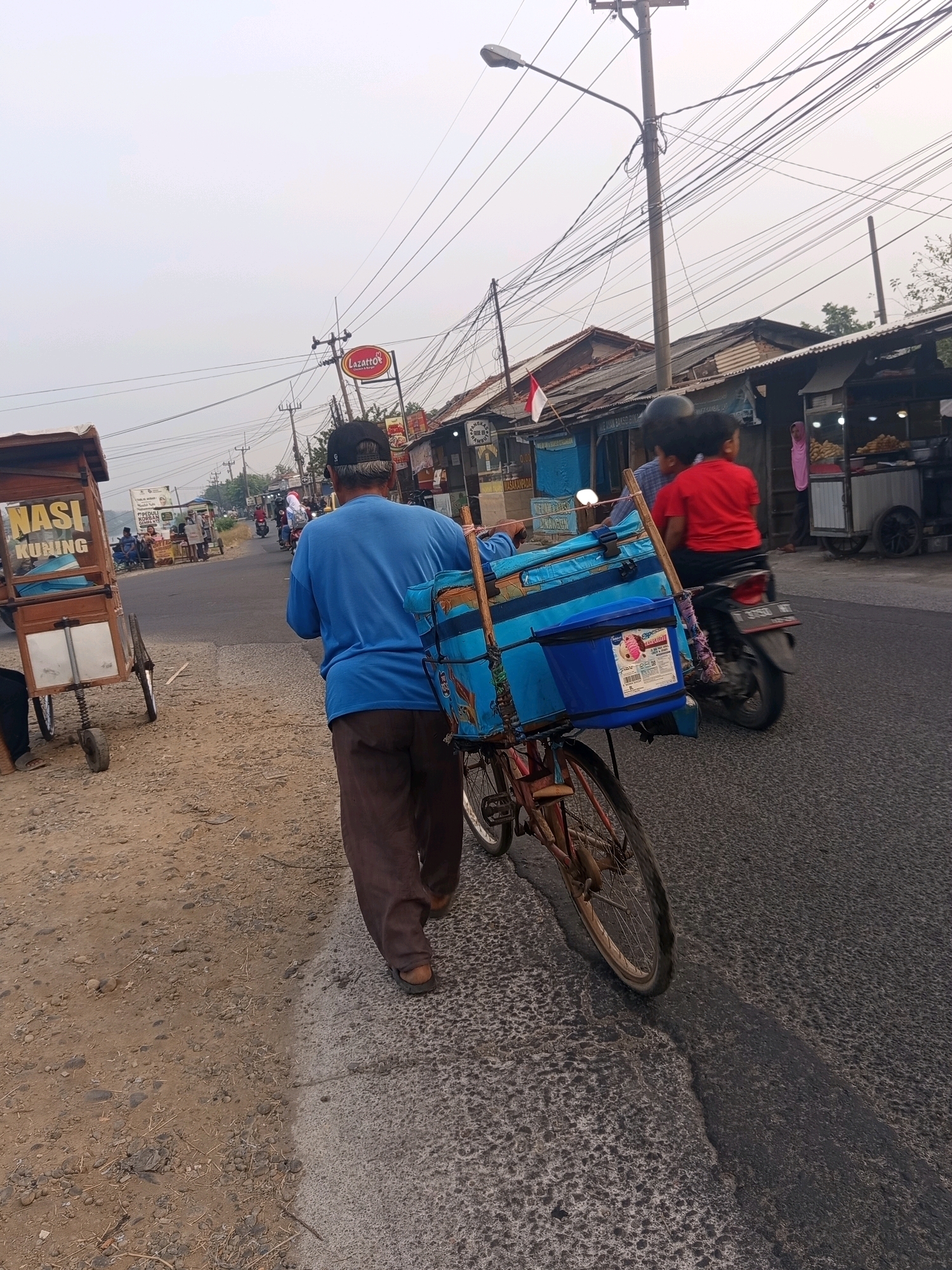 In Karawang, Indonesia, a street scene unfolds. A man wearing a blue shirt and brown pants walks away from the camera, pushing a bicycle loaded with blue insulated containers. A smaller blue container with a label depicting ice cream is attached to the front of the bicycle. The bicycle has a red frame and a spoked wheel visible. The man is also wearing a black cap.
To the left of the man, a food cart with the words "NASI KUNING" (Yellow Rice) printed on it is parked on a dusty roadside. The cart has a wooden frame and two wheels. Further down the road, a motorcycle is seen with two passengers, a child in a red shirt and another individual wearing a helmet.
The environment consists of a paved road, a dusty shoulder, and a row of buildings with corrugated metal roofs lining the right side of the road. Overhead, a complex network of electrical wires is strung between utility poles, and a streetlamp is visible. The sky is overcast, suggesting a possibly hazy or cloudy day. The overall impression is that of a typical street scene in a developing urban or semi-urban area.