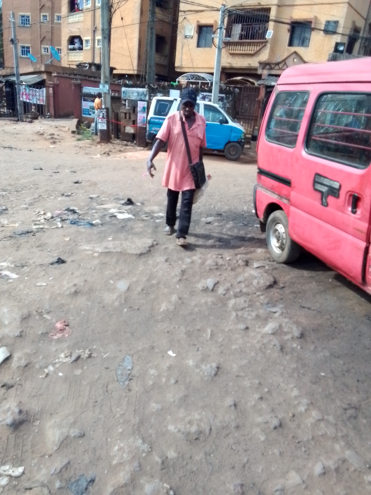 The image depicts a street scene in Onitsha, Nigeria, characterized by a deteriorated, unpaved road littered with debris. A man wearing a pink shirt, dark pants, and a black cap walks across the road, carrying a dark bag slung across his shoulder. A pink van is parked on the right side of the frame. In the background, buildings of varied heights and colors are visible, as well as a blue and white vehicle. A sign is posted near a building with an orange structure.