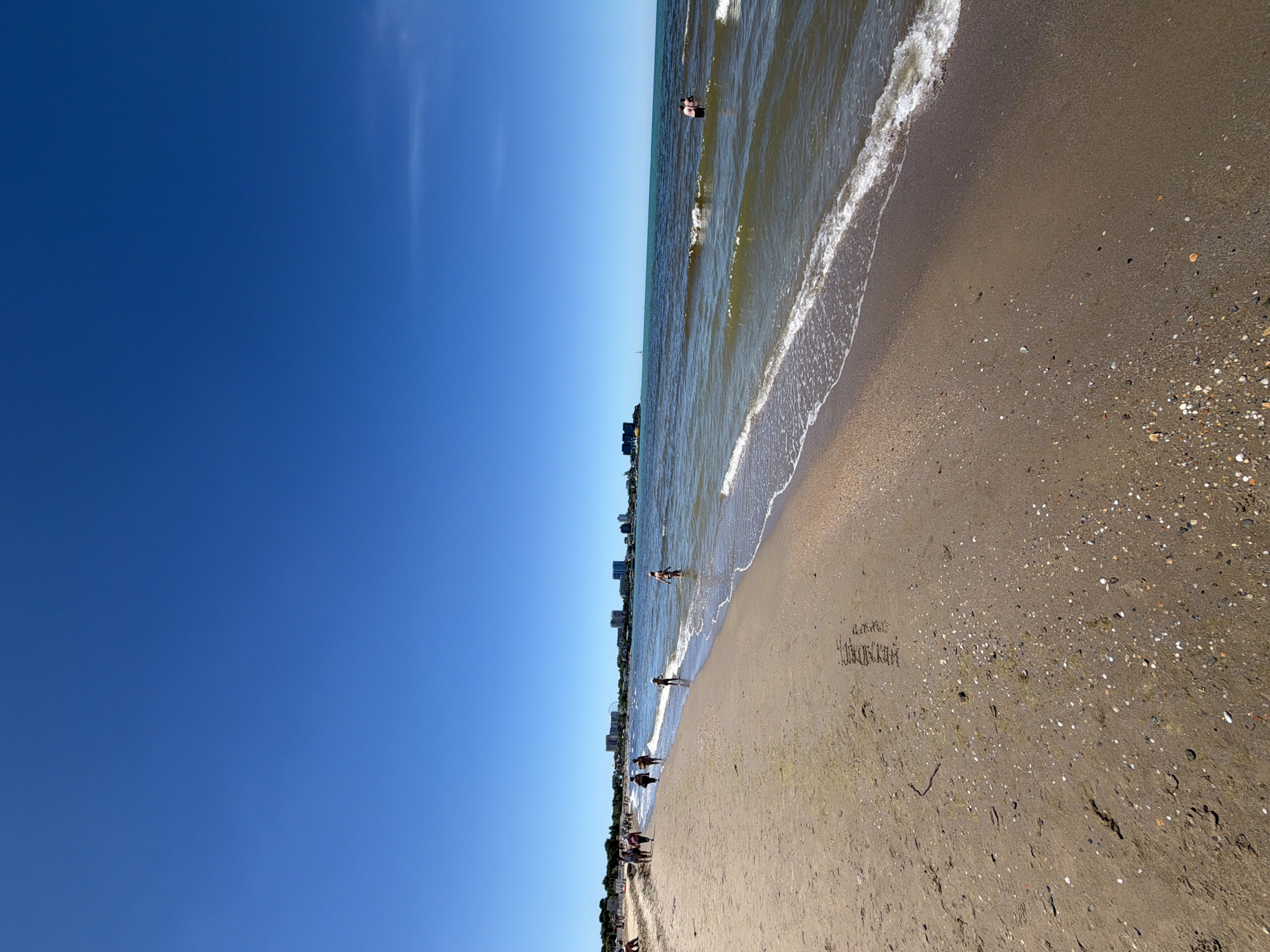 The image displays a vibrant beach scene, viewed in its correct upright orientation, under a bright, clear sky, likely in Anapa, Russia, as indicated by the context and visible text. The upper portion of the frame is dominated by an expansive, cloudless blue sky, with only a faint wisp of white cloud visible towards the top right.
Below the vast sky lies the calm, greenish-blue sea, characterized by gentle waves breaking into white foam along the shoreline. Several people are visible in the water; one individual in dark swim attire wades in the shallow waves in the mid-ground, while another person is partially submerged further out. In the distance, along the horizon where the sea meets the land, a line of buildings suggests a coastal town or resort.
The lower half of the image showcases the sandy beach, which is a light brown color. Its surface is speckled with numerous small white, gray, and dark pebbles and shell fragments, particularly denser towards the water's edge and the foreground. Further along the beach to the right, a small group of people are walking or standing, and some indistinct beach furniture can be vaguely discerned.
A notable detail is the handwritten text etched into the sand in the lower right quadrant. Written in Cyrillic, it reads "Я был в" (I was in) on the first line, followed by "Анапа" (Anapa) on the second line, explicitly confirming the location. The overall atmosphere conveys a peaceful and enjoyable summer day at the seaside.