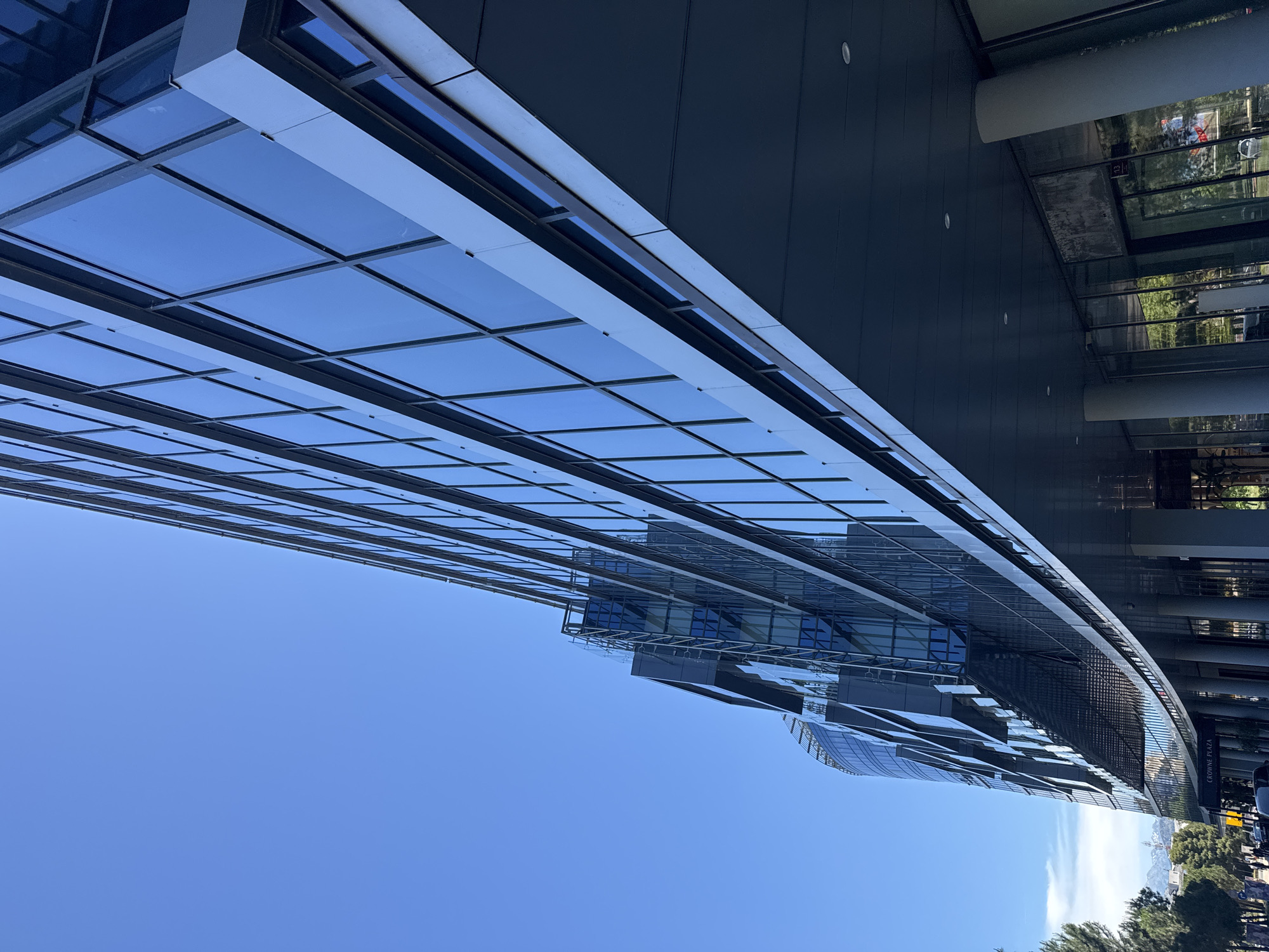 The image shows a modern glass building, likely an office complex, under a clear blue sky. The building's facade is primarily composed of rectangular glass panels arranged in a grid pattern. It appears to be located in Podgorica, Montenegro, based on the hotel brand and architectural style. The image captures an upward angle view of the building, emphasizing its height and architectural details. Some foliage is visible at the base of the building, suggesting an urban environment with landscaping.