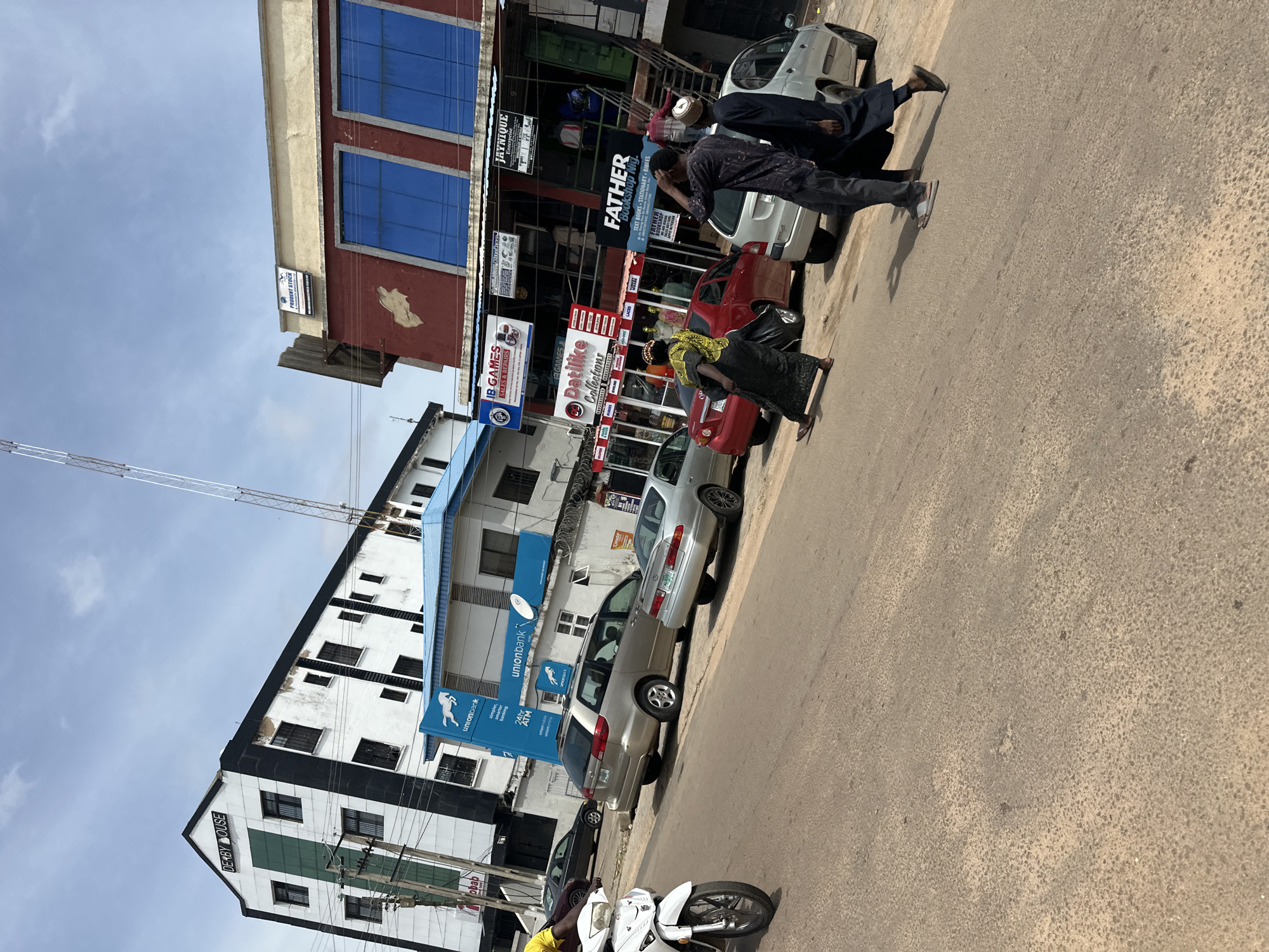 A street scene in Jos, Nigeria, shows a white motorcycle in the foreground to the left, facing right. Several cars are parked and driving on the asphalt road. Buildings line the street, including "DERBY HOUSE" on a white and black building with green accents, and another building with a red facade and blue windows. Signs for "Union Bank," "IB GAMES," "Datilike Collections," and "FATHER Bookshop NG" are visible. Three individuals are walking across the road, two men in dark clothing and a woman in a vibrant yellow and black dress. The sky is partly cloudy.