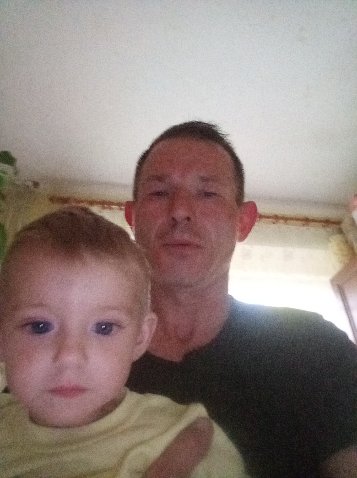 The image shows a close-up, selfie-style photograph of two individuals, an adult male and a young child, both appearing to be indoors. The adult male is positioned behind and to the right of the child, with his face partially visible. He has short, dark hair and is wearing a dark t-shirt. The child, in the foreground and larger in the frame, has light-colored hair and blue eyes, and is wearing a light-colored t-shirt. The background appears to be a room with a white ceiling and a window with curtains. No specific landmarks or environmental cues suggest the city of Tapa, Estonia.