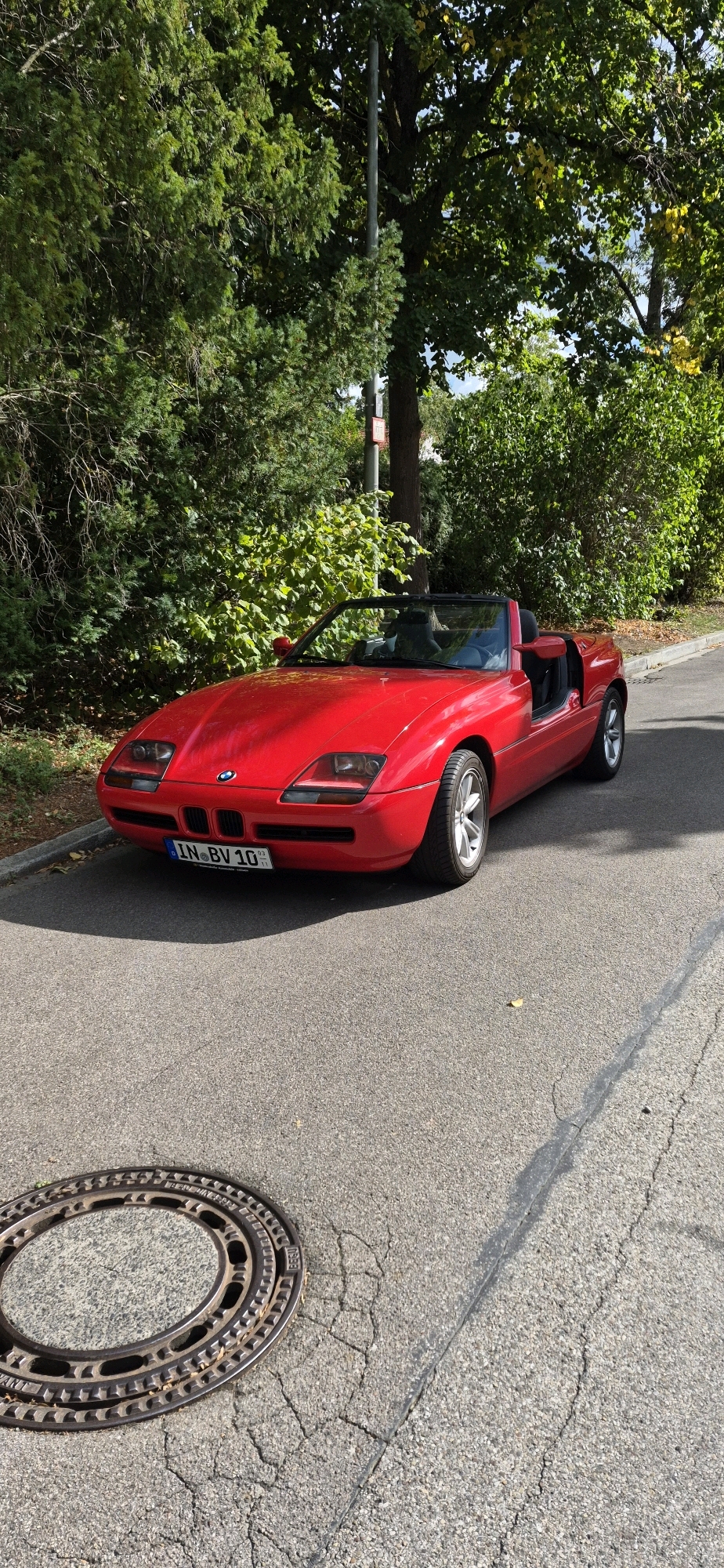 A red BMW Z1 convertible is parked on an asphalt street, angled towards the camera. The car has its doors open, revealing black leather seats. The front of the car features the distinctive BMW kidney grille and pop-up headlights. A license plate reads "IN BV 10". The car is situated on a street lined with lush green trees and bushes. In the foreground, a circular manhole cover is visible on the asphalt. The scene appears to be a quiet residential street or park area within Ingolstadt, Germany, indicated by the license plate. The lighting suggests it is daytime.