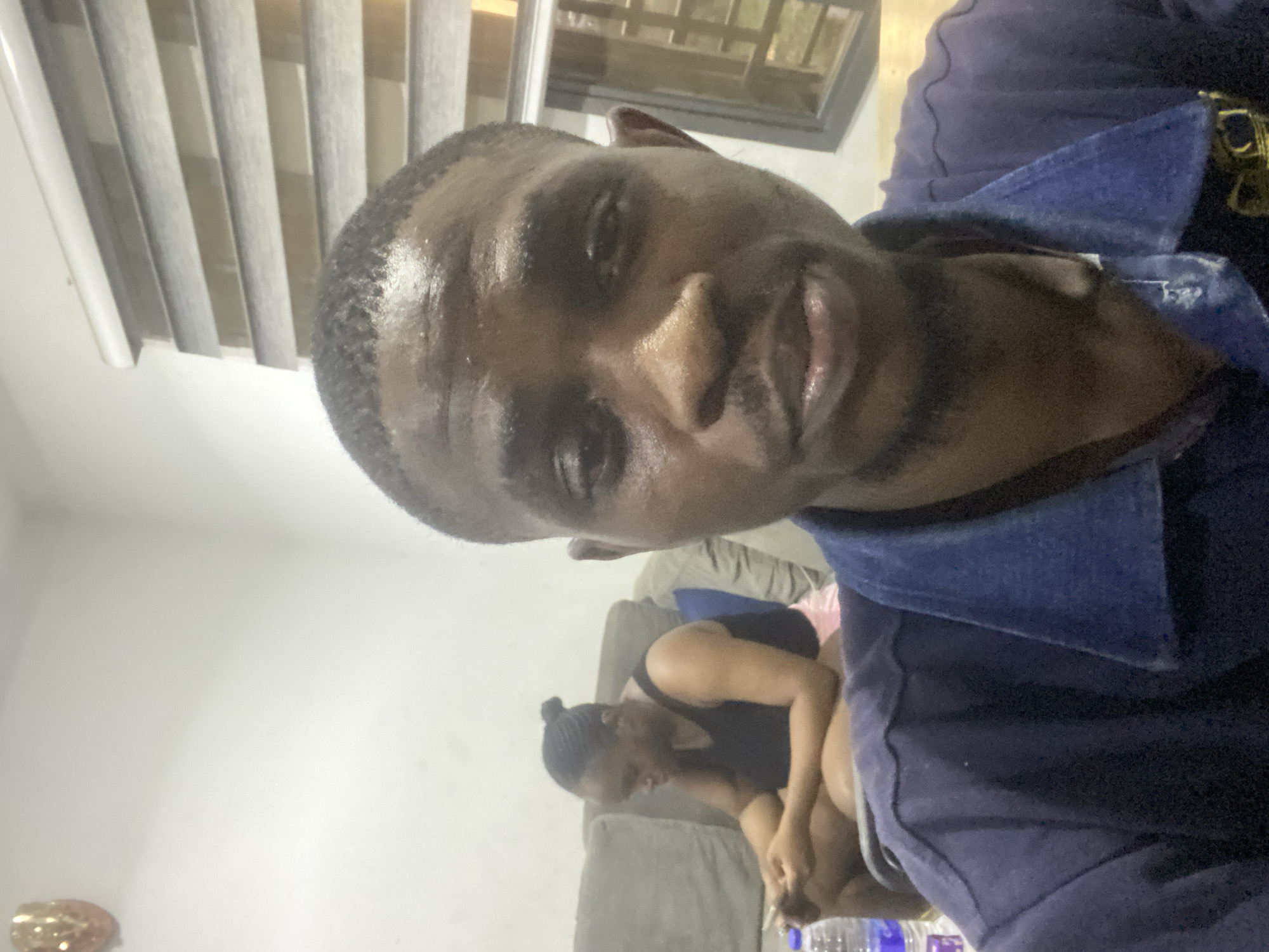 This vertically oriented image is an indoor selfie featuring two individuals. The main subject, a man with dark skin, short dark hair, and light facial hair, dominates the foreground. He is wearing a dark blue or navy collared shirt and looks directly at the camera with a neutral to slightly smiling expression. His face appears somewhat shiny, possibly from lighting.
In the mid-ground, seated on a gray sofa, is a second person, likely a woman, with dark hair pulled into a bun. She is wearing a dark-colored top and appears to be leaning forward, focused on an object in her hands, with her face partially obscured.
The setting is an indoor residential space, possibly a living room, with light-colored walls. Artificial lighting creates a dim, warm atmosphere, suggesting it is nighttime or evening. Details include vertical window blinds in the top left, a dark-framed window or door section in the upper right, and several clear plastic water bottles on the floor near the sofa. A gold-toned, circular decorative item is visible on a surface in the bottom left corner. No visible text is present. The scene depicts a relaxed environment in Abuja, Nigeria.
