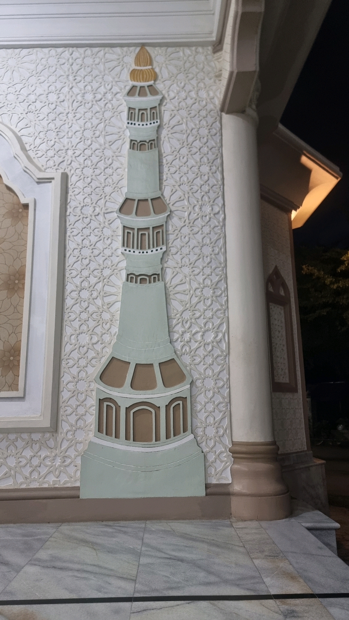 The image shows an exterior view of a building in Lhokseumawe, Indonesia, at night. A decorative minaret, painted in muted green and brown, is prominently featured on the left side of the frame, attached to a wall with intricate white geometric patterns. To the right of the minaret, a large, light-colored pillar supports an overhang. The ground in the foreground is paved with marble tiles. The background is dark, with hints of trees and a dimly lit structure visible on the right. The overall impression is one of a religious or cultural building, possibly a mosque, illuminated by artificial light.