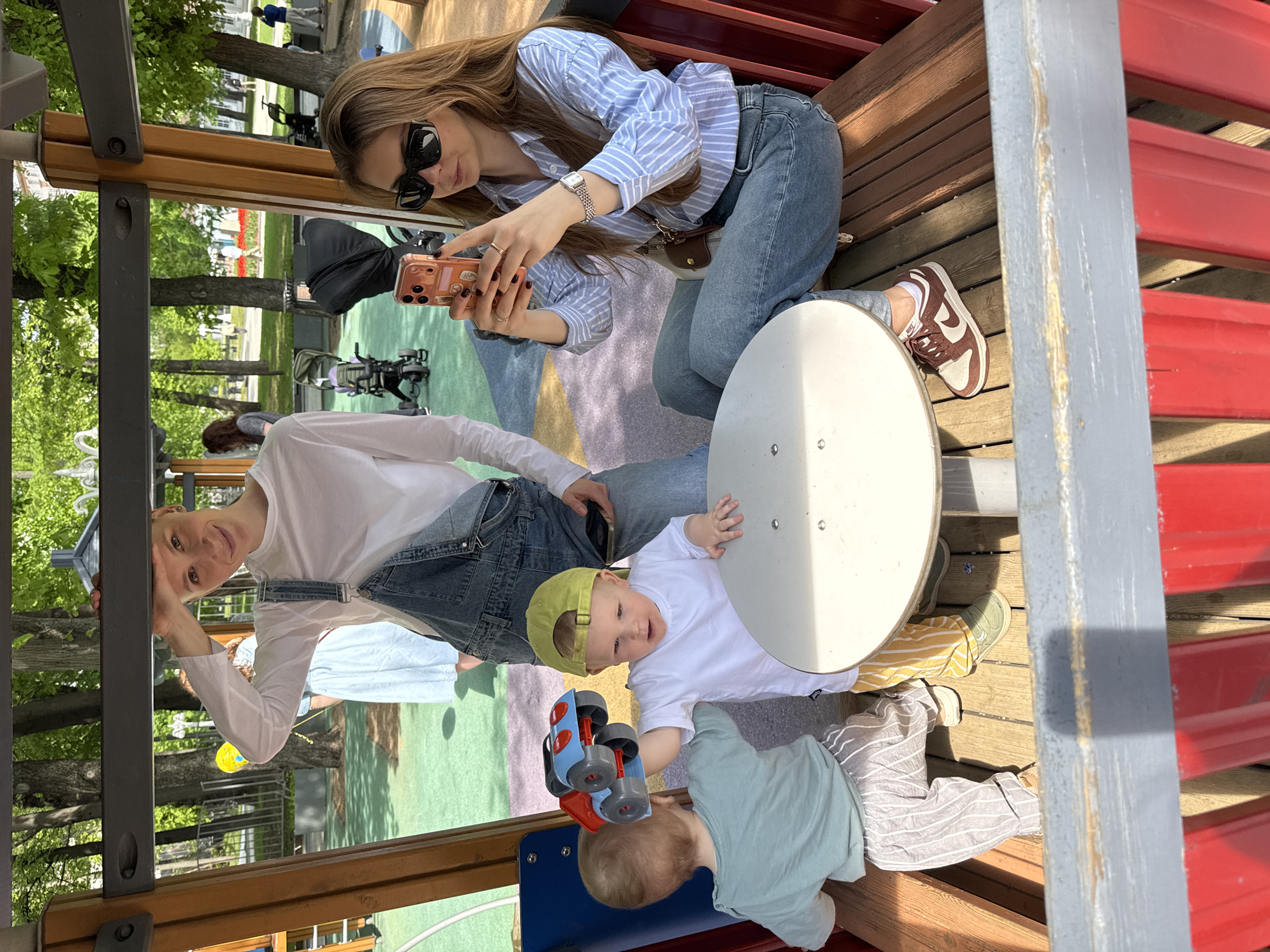 The image captures a scene at a playground, possibly in Moscow, Russia, evidenced by the architecture and layout characteristic of the city's urban spaces. A woman with long brown hair, wearing a striped blue shirt, blue jeans, and sunglasses, is taking a photo with her smartphone. She is seated on a wooden platform. An adult in white shirt and denim overalls and two infants (one in striped pants and the other with a green hat, white shirt and striped pants) are at the playground. The two infants are in close proximity to each other; one is holding a toy. The setting appears to be sunny with green trees visible in the background.