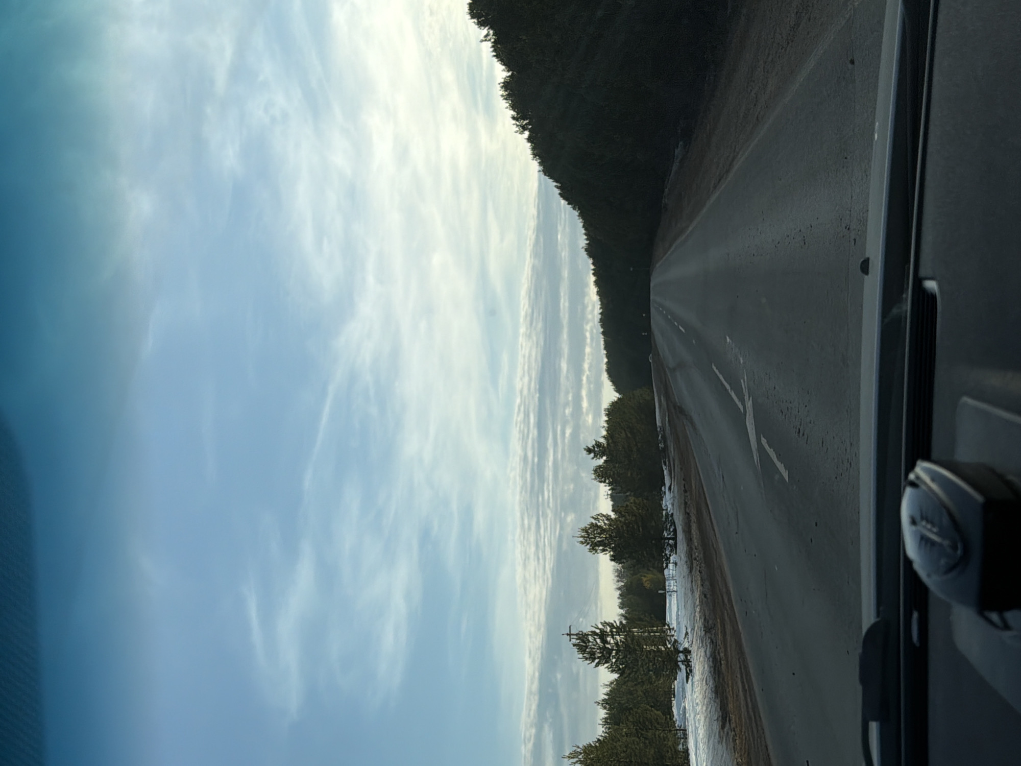 The image depicts a rural road scene, likely taken from the interior of a moving vehicle, near the city of Фоки, Russia. In the foreground, the upper portion of a dark dashboard is visible, centrally featuring a black electronic device, possibly a radar detector, mounted on its surface. Above the dashboard, the windshield frames a two-lane asphalt road that extends into the distance, exhibiting a dull, possibly damp or dirty, surface. A faded white arrow road marking is present on the left lane.
To the left of the road, the ground is covered with patches of melting snow, interspersed with bare earth and scattered evergreen trees, predominantly pines. A wooden utility pole with crossarms and insulators stands upright among these trees. To the right of the road, a dense, dark green forest lines the roadside, suggesting a slightly elevated terrain. The sky occupies the upper two-thirds of the image, displaying a gradient from light blue at the horizon to a deeper blue-green overhead. Scattered, wispy white clouds are visible across the expanse of the sky. No individuals or other vehicles are visible within the frame.