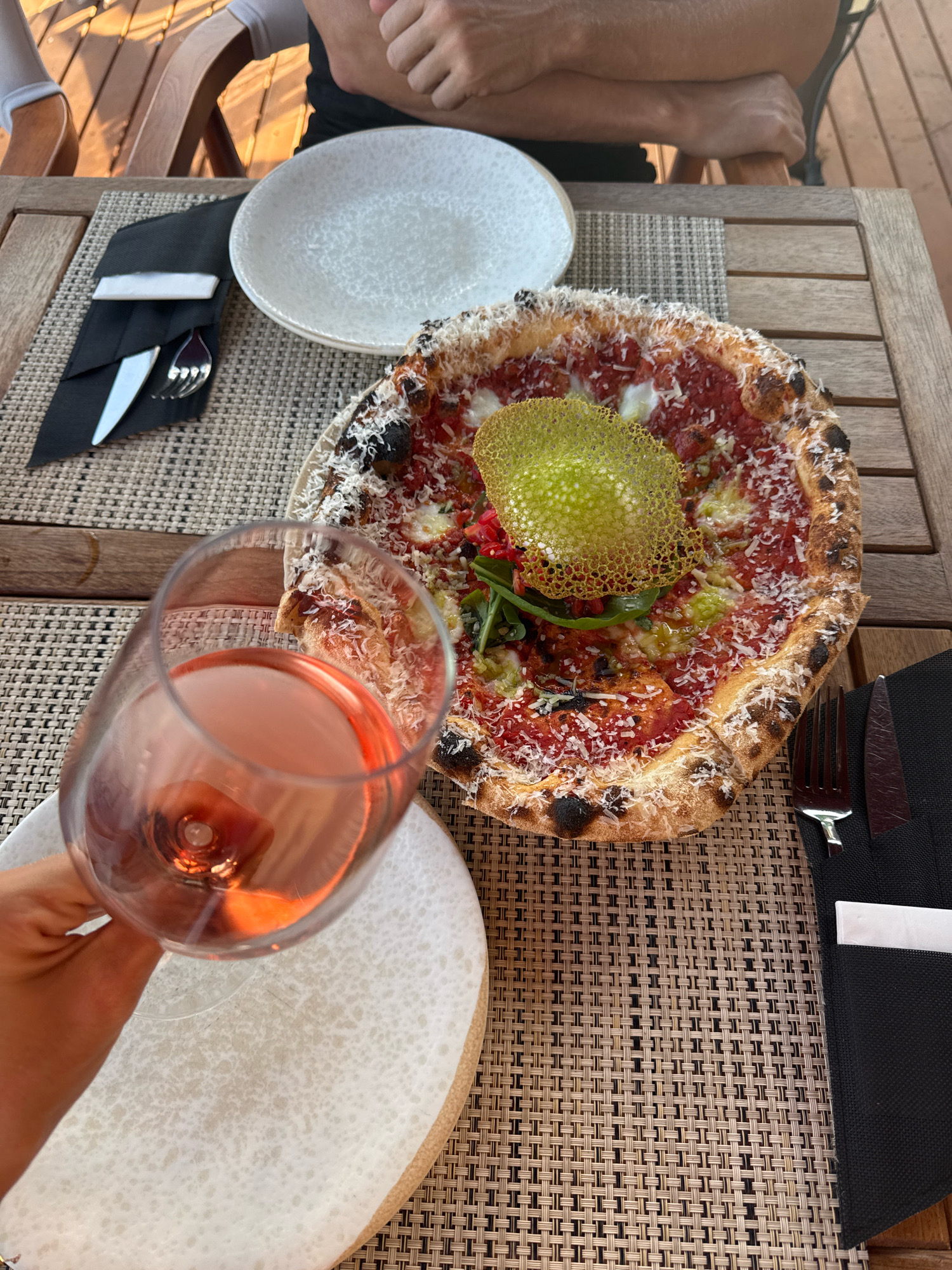 An overhead shot shows a table setting at an outdoor restaurant in Konyaaltı, Turkey. A pizza with a golden crisp topping and grated cheese sits on a woven placemat. A glass of rosé wine is held in the foreground, obscuring a white plate. The table is made of wood, and other diners' arms are visible in the background.