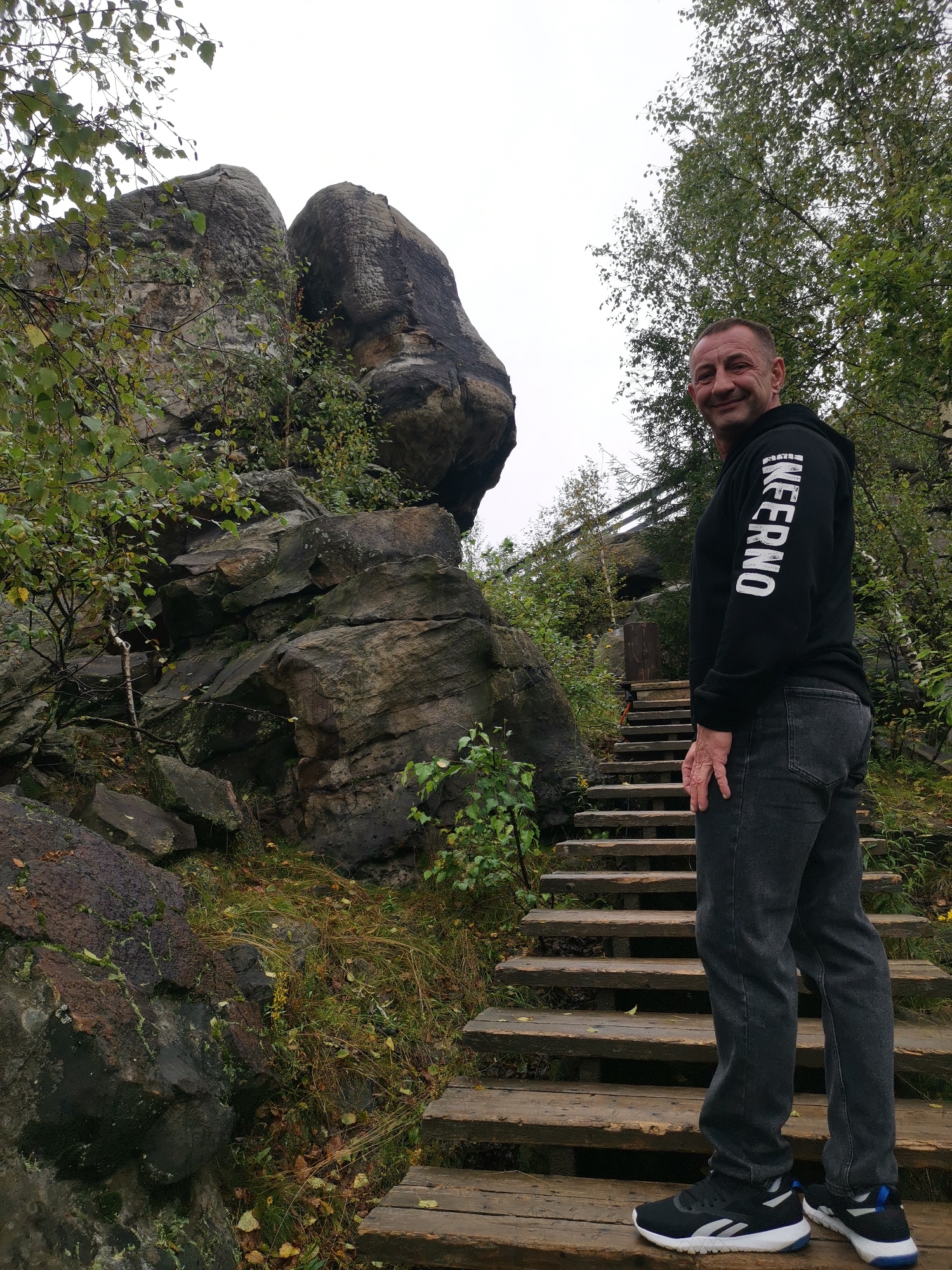 In Gubakha, Russia, a man stands on a wooden staircase next to large rock formations. He is wearing a black hoodie with white lettering on the sleeve and dark grey jeans, and black and white athletic shoes. The man is smiling and looking at the camera. The staircase is made of weathered wooden planks and leads upwards. The rock formations are rough and weathered, with moss and vegetation growing on them. The background consists of trees and a cloudy sky.