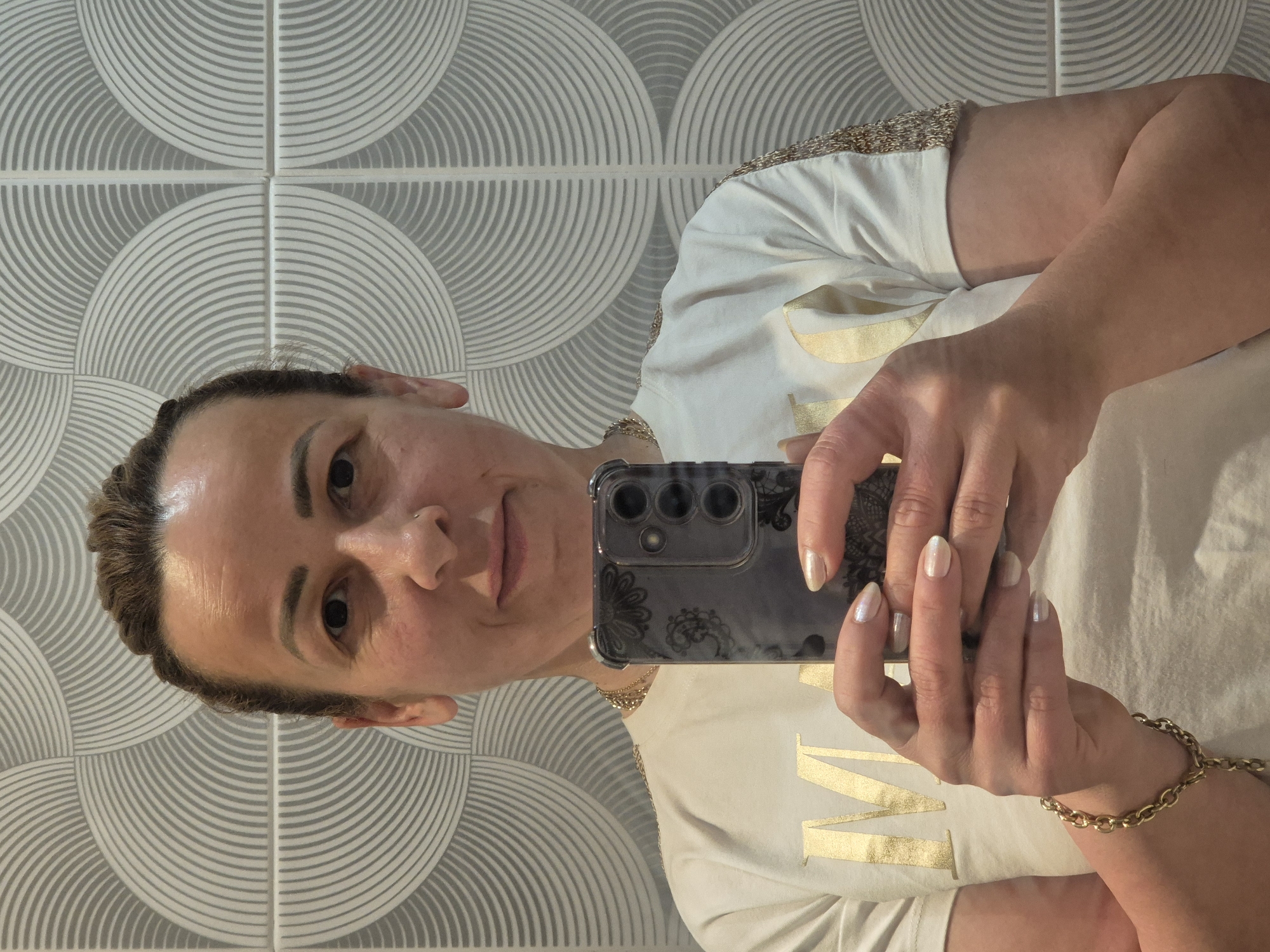 A woman is taking a selfie in front of a tiled wall. She is holding a dark-colored smartphone in her hands, which are positioned in the lower center of the frame. The woman has her hair pulled back and is wearing a white t-shirt with gold lettering and gold chain detailing on the sleeves. She is also wearing a gold chain necklace and a gold bracelet on her left wrist. The wall behind her features a repeating geometric pattern in shades of gray and white. The lighting appears to be artificial and illuminates the woman's face and the foreground.