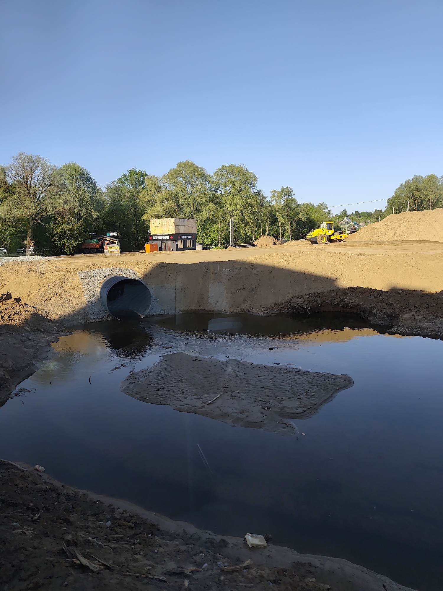 The image depicts a construction site in Oktyabr'skii, Russia. A large concrete culvert, presumably for drainage, is visible in the center of the frame, with one end submerged in a body of water, reflecting the sky. The culvert is embedded in a mound of tan-colored sand and dirt. Various pieces of construction equipment are present, including a yellow road roller on top of the mound and a dump truck parked on the elevated surface behind the mound. A structure with Cyrillic writing is also seen in the background. Trees line the horizon under a clear, light blue sky. The foreground is dominated by the body of water, showing muddy banks and a sandbar.