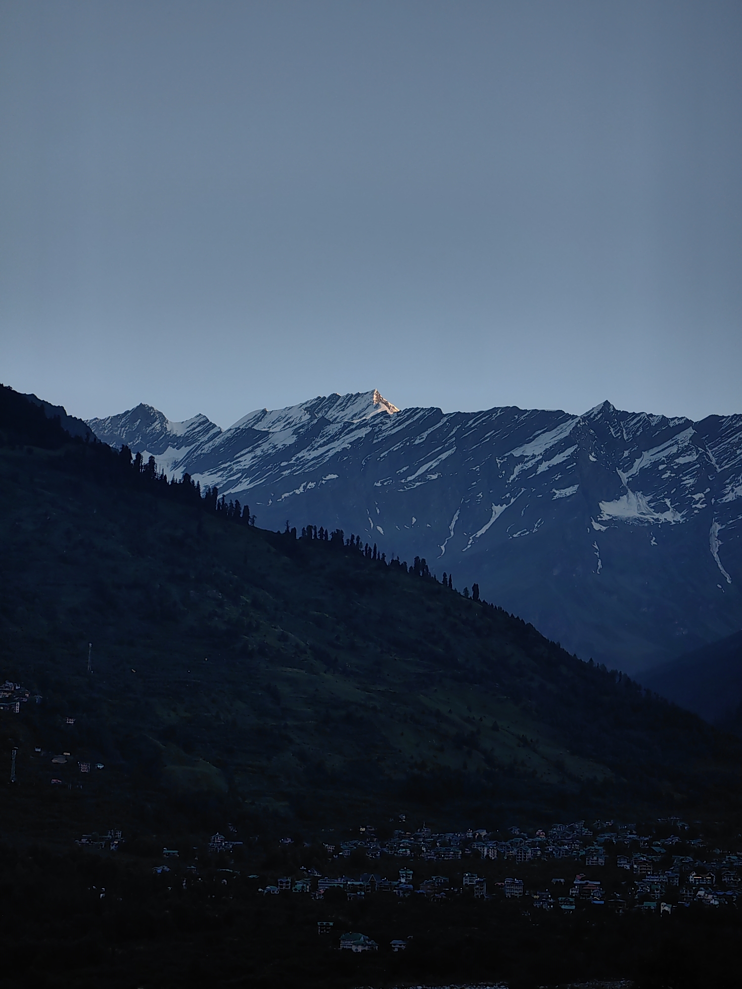 The image depicts a mountainous landscape at twilight, likely in or near Vashisht, India. The sky is a deep, clear blue, fading to a lighter shade near the horizon. In the foreground, a dark, tree-covered mountainside slopes down towards a cluster of illuminated buildings, suggesting a village or town. The buildings are small and appear to be a mix of houses and possibly guesthouses, with some lights on, indicating the presence of inhabitants.
Further back, a majestic range of snow-capped mountains dominates the background. The peaks are sharp and angular, with patches of snow clinging to their upper slopes. The setting sun casts a warm, golden glow on one of the highest peaks, creating a striking contrast against the cool blue of the sky and the dark mountains. A line of dark, conical trees is visible along the ridge of the nearer mountain, silhouetted against the sky. The overall impression is one of serene natural beauty, with human settlement nestled harmoniously at the foot of the imposing Himalayas.