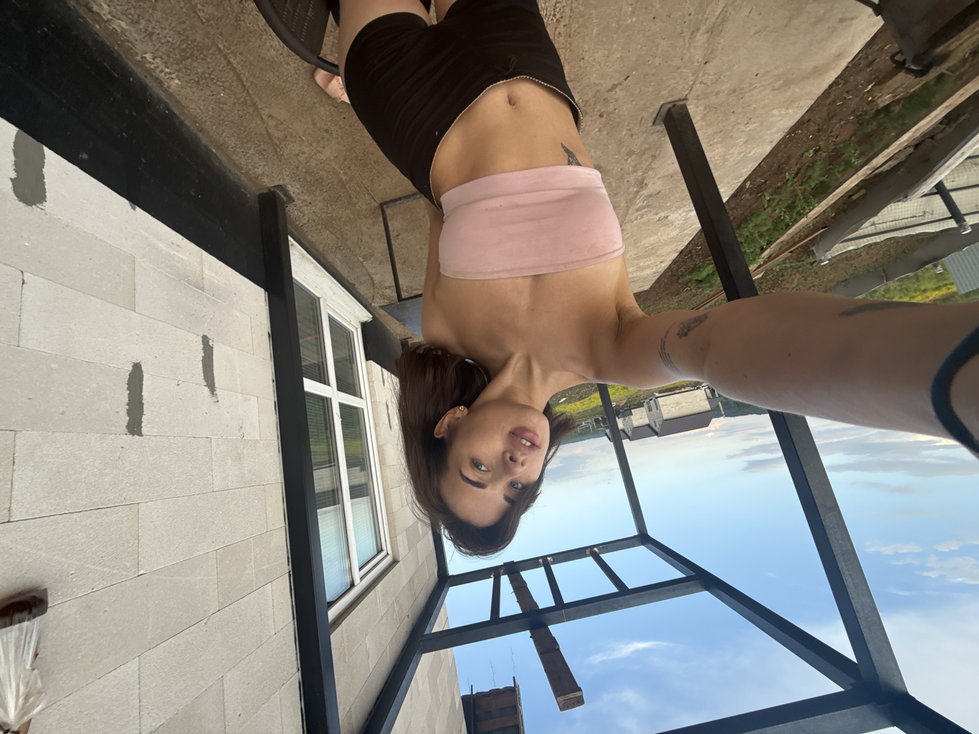A woman with dark hair and fair skin poses for a selfie, her face positioned towards the camera. She is wearing black shorts and a pink bandeau top. The photo is taken from a low angle, showing a building under construction behind her. The structure appears to be metal framing against a cloudy sky. The location is Лобаново, Russia.