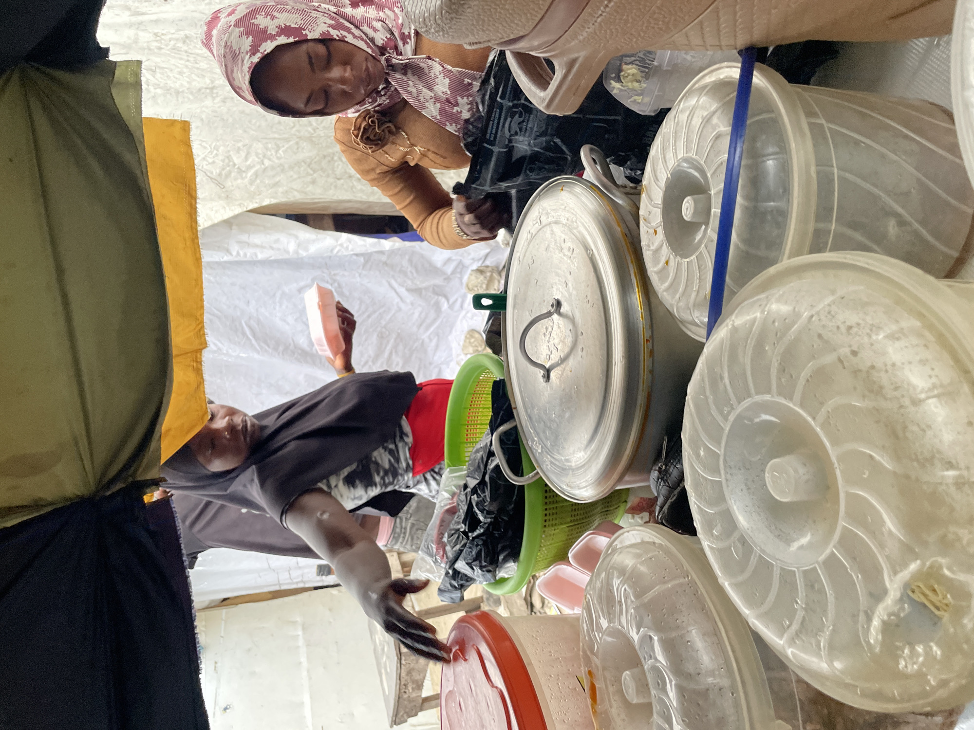 In Ilado, Nigeria, this photograph captures a busy scene at what appears to be a market stall, possibly selling food. Two women, dressed in traditional Muslim attire with headscarves, are engaged in their activities. The woman on the left, wearing a black hijab and a black and white patterned top with a red skirt, is reaching towards a stack of large, clear plastic containers with red and transparent lids. Her left hand is extended, and she appears to be either organizing or selecting something. Beside her, a green plastic basket overflows with black plastic bags, suggesting packaged goods. In front of these containers, a large, silver pot with a lid and handle is visible.
The woman on the right, her head covered with a pink and white patterned hijab and wearing a light brown cardigan adorned with a fabric flower, is looking down. Her hands are occupied with a black plastic bag, possibly sorting its contents. Behind her, another large, light brown container or cooler is visible. The stall is covered by a makeshift awning made of black and olive green fabric, with a yellow panel hanging below. In the background, white sheeting creates a backdrop, and a wooden stool is partially visible. The overall impression is one of active commerce and daily life in a Nigerian marketplace.