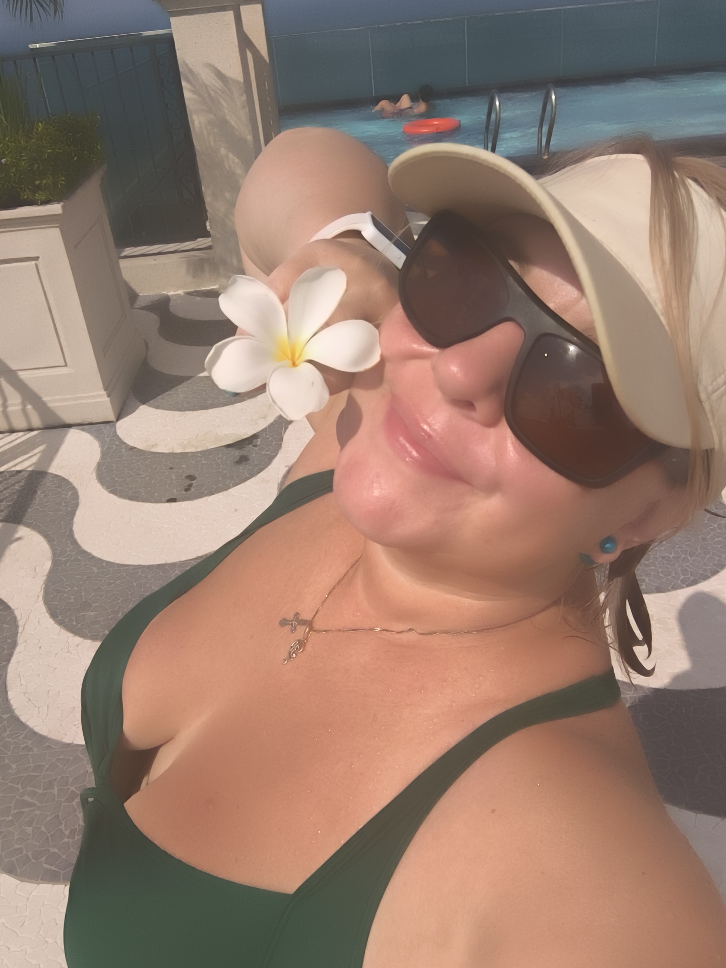 A woman is lying on a lounge chair by a pool, holding a white flower with a yellow center to her face. She is wearing a green bikini top, a beige visor, and sunglasses. There are two people in the pool behind her, one of whom is wearing an orange flotation device. The background includes a tiled patio with a wavy pattern, a planter with greenery, and a fence. The setting appears to be a resort or hotel in Phú Quốc, Vietnam.