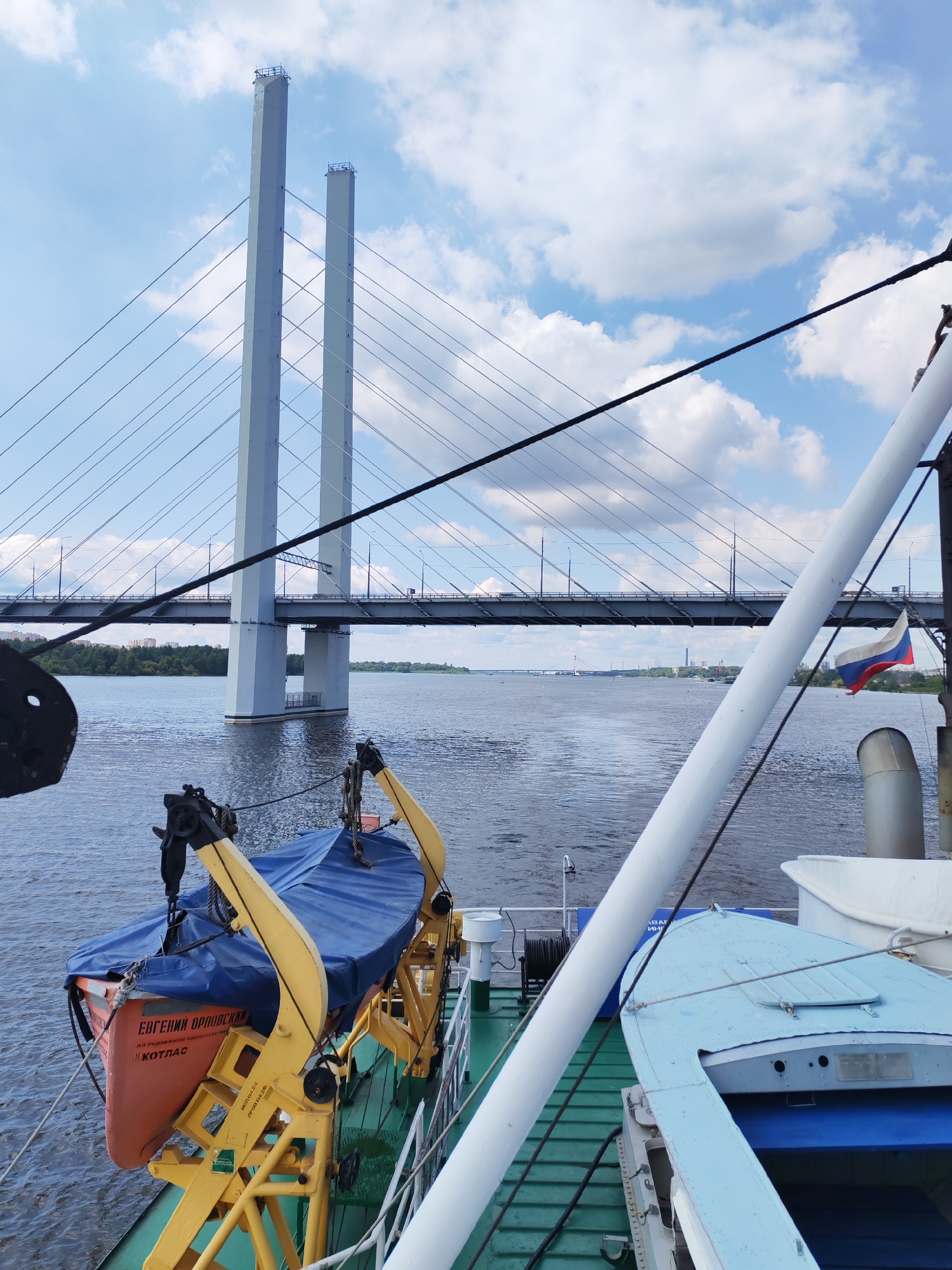 The image shows a view from a ship on a body of water, likely a river. In the foreground, parts of the ship's deck are visible, including a life raft with a blue cover, a yellow davit, and a light blue boat. Further in the distance, a large cable-stayed bridge spans the water. The sky is partly cloudy. A Russian flag is visible on the right side of the image, attached to a pole. The water is choppy, and the overall scene suggests a cloudy but otherwise clear day.