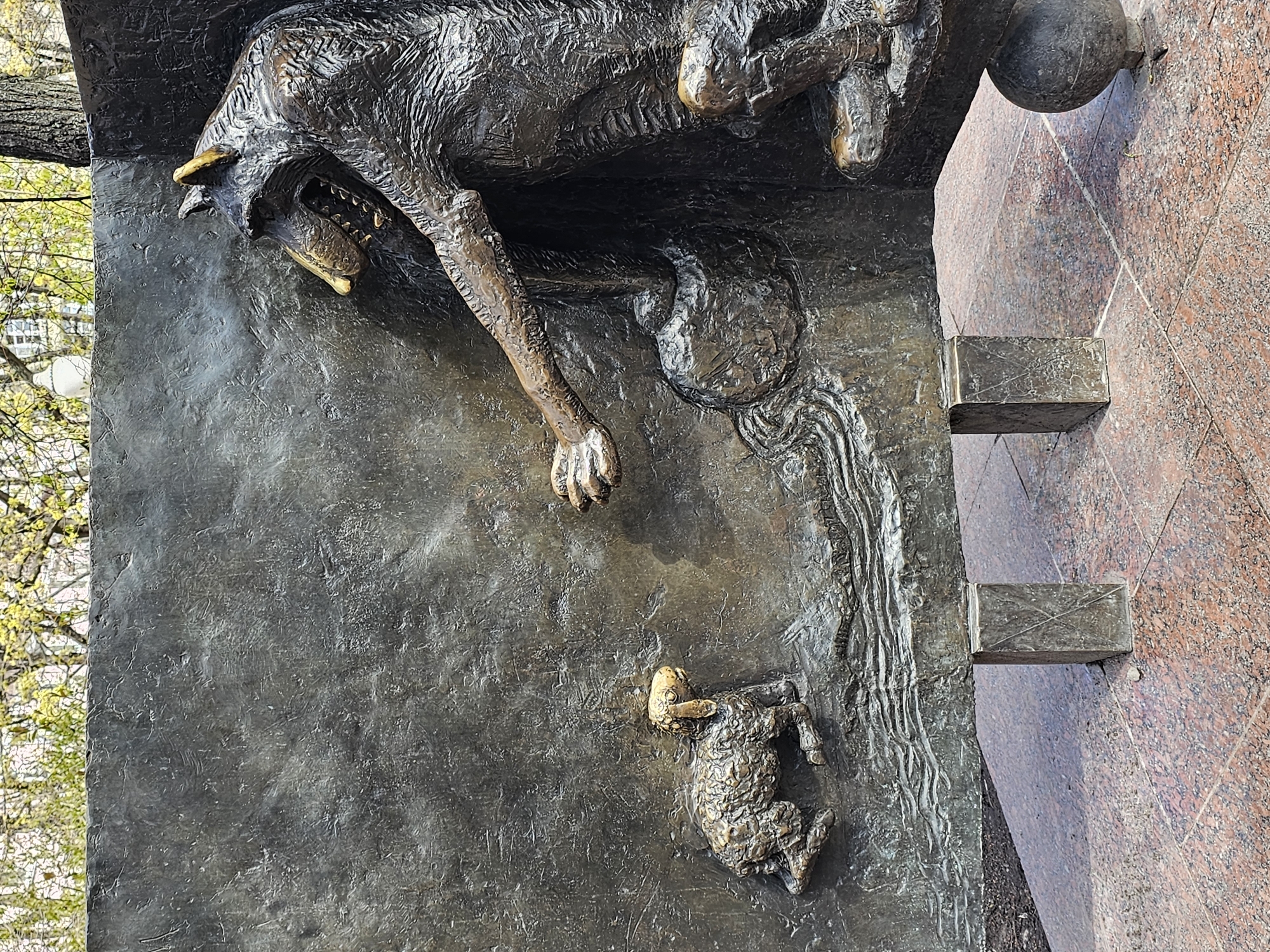 This is a close-up of a bronze relief sculpture depicting a wolf with a lamb. The wolf, rendered with visible teeth and claws, appears to be suspended or attacking. Below it, a small lamb is depicted. A design element reminiscent of flowing water connects the two figures. The sculpture is mounted on a red granite base, and a tree with budding leaves is visible in the background. This sculpture is located in Moscow, Russia. It is likely a fragment of a larger monument.