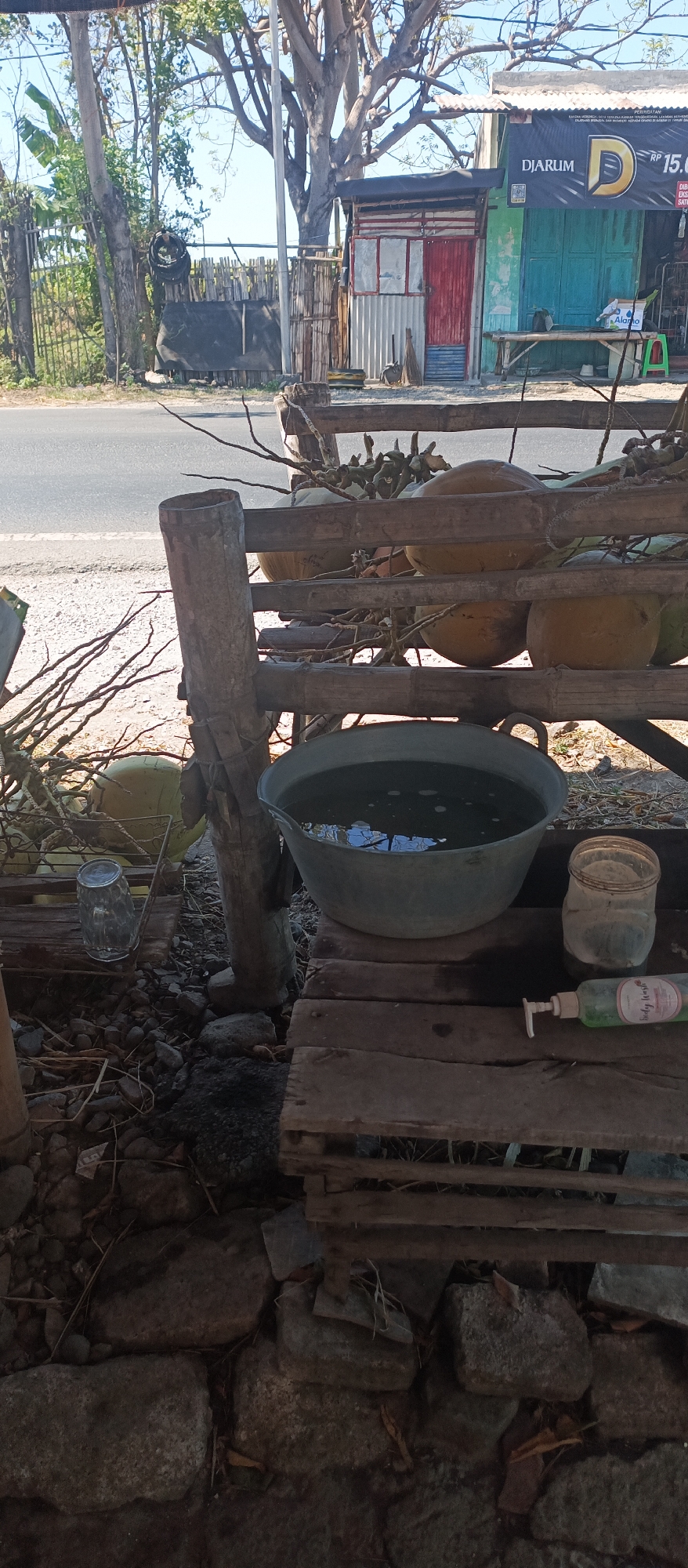The image captures a rustic outdoor scene near a road, possibly in Jatisari, Indonesia. In the foreground, a wooden structure, appearing weathered and assembled from rough-hewn timber and bamboo, supports several objects. A large, galvanized metal basin filled with water sits prominently on the structure. To its right, a clear glass jar, an empty plastic container, and a bottle of liquid soap with a pump dispenser are placed on a wooden surface. The background reveals a roadside shop with a corrugated metal facade painted in green and red, adorned with a banner that reads "DJARUM". A wooden bench is visible in front of the shop, and further back, a fence made of bamboo poles encloses an area with trees and vegetation. The ground is covered with stones and dry leaves, suggesting a rural or semi-urban environment. The overall impression is one of a simple, perhaps temporary, setup for washing or personal hygiene in an outdoor setting.