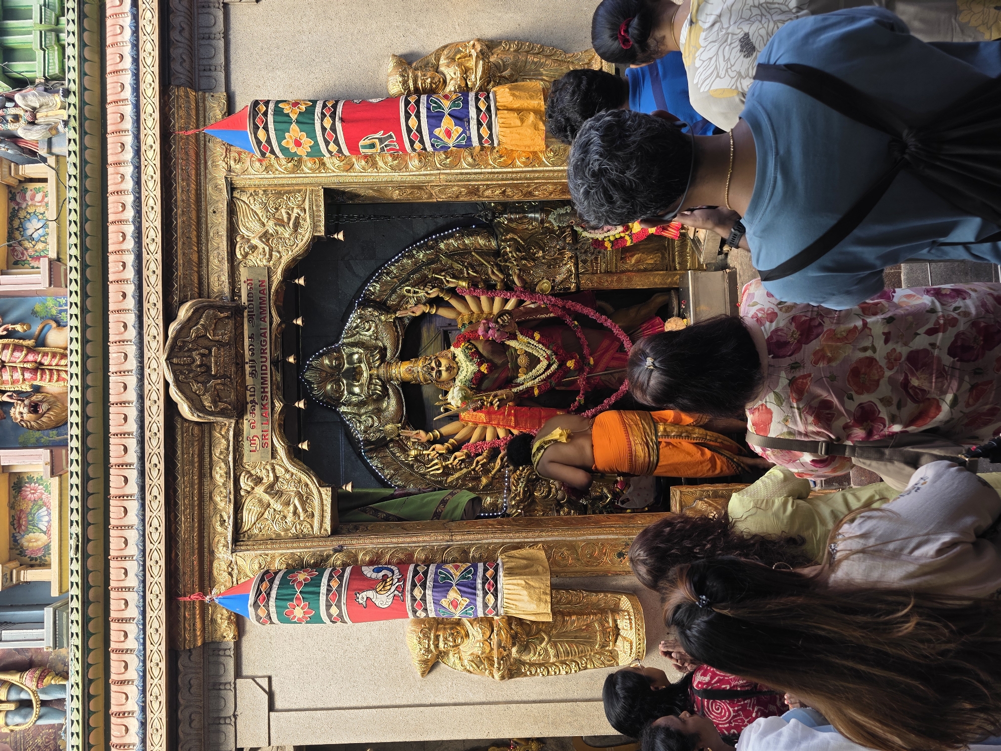 In Singapore, a group of people are gathered in front of a Hindu temple shrine, likely within the city. The shrine is elaborately decorated with gold-colored statues and ornate carvings. A large statue of a multi-armed deity, possibly Durga, is the central focus, adorned with garlands. A man in an orange garment stands facing the deity, with his back to the viewer, seemingly in prayer or performing a ritual. A line of individuals, mostly women, are positioned behind him, observing the shrine. The architecture includes tall, colorful cylindrical decorations flanking the entrance. The scene suggests a religious gathering or visit to a place of worship.