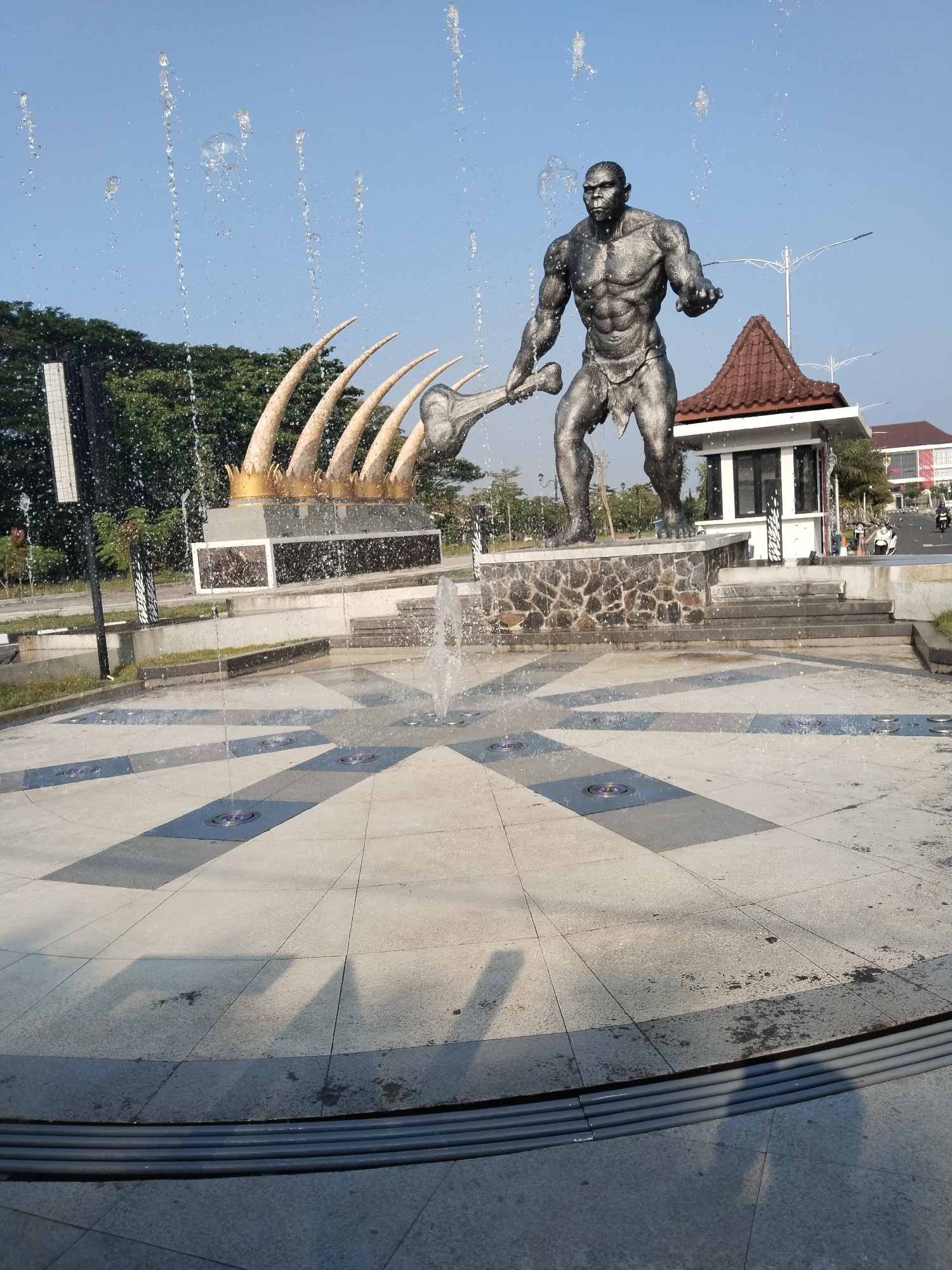 The image displays a public square or roundabout featuring a large, prominent statue of a muscular, primitive human-like figure, often referred to as an "ape-man" or early human, holding a substantial club-like weapon. This bronze-colored statue stands on a stone-faced pedestal within a circular fountain. Numerous jets of water spray upwards and outwards from the fountain, creating a dynamic effect with water droplets visible against the clear blue sky.
Behind the main statue, a second monument is situated, characterized by several large, curved, ivory-colored structures resembling tusks or ribs, each adorned with a golden crown-like detail on top. This monument rests on a multi-layered base of white and dark panels.
The background includes a dense line of green trees to the left, and to the right, a small building with a distinctive brown, traditional Javanese-style roof. Beyond this structure, a road is visible with streetlights and a few motorcycles.
The scene is captured in bright daylight under a clear blue sky, casting distinct shadows on the light and dark grey tiled pattern of the fountain's ground. The active fountain and the themes of early human figures and bone-like structures suggest a location with a focus on prehistoric heritage, consistent with Sragen Kulon, Indonesia. No explicit text or specific human activity is discernible in the immediate foreground, only distant traffic.
