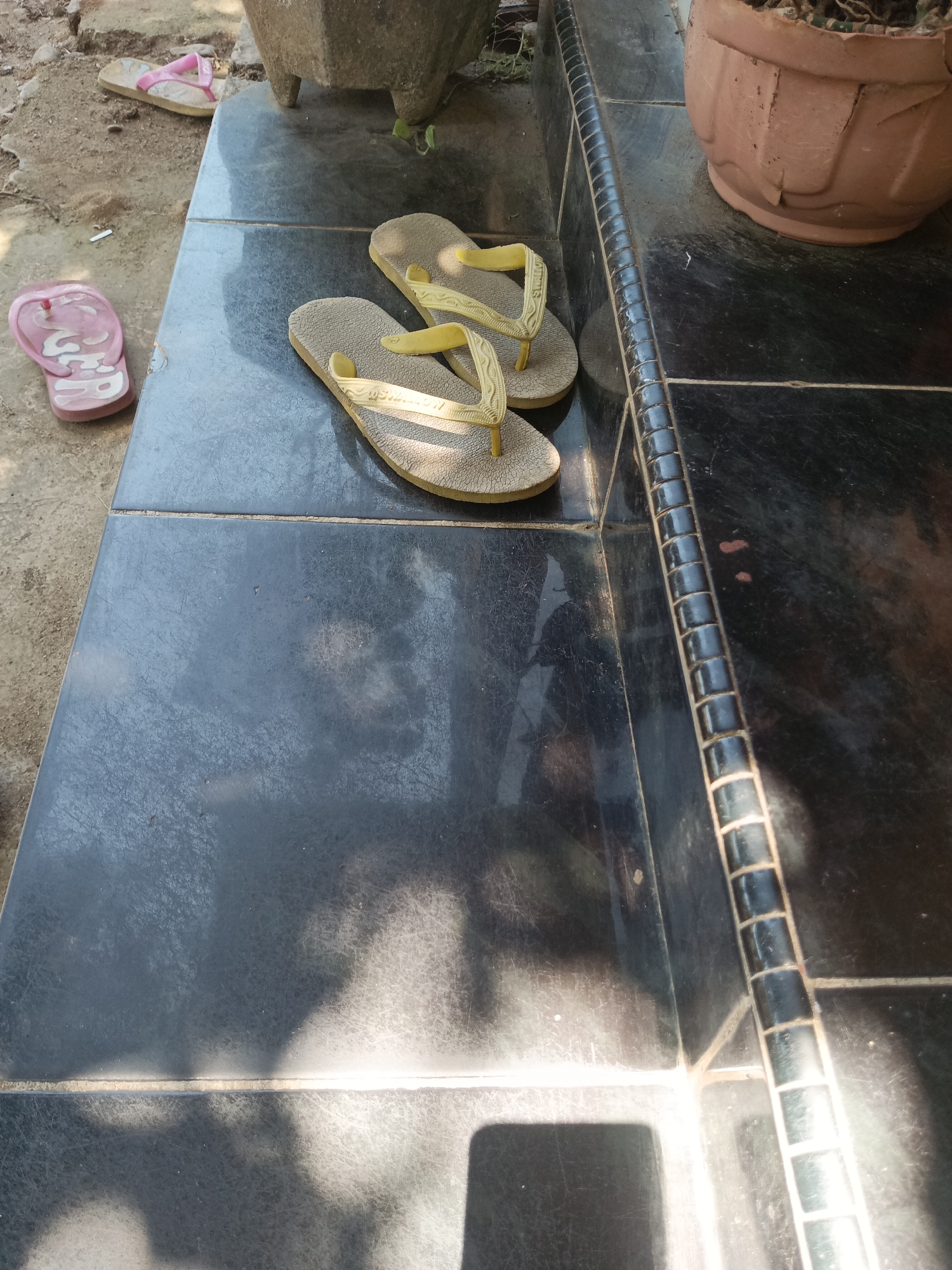 The image depicts a pair of light yellow flip-flops resting on a dark, glossy tiled surface. To the left, there are two pink flip-flops on a rough, sandy ground. Behind the yellow flip-flops, a portion of a large, weathered terracotta pot with a plant is visible. The scene appears to be outdoors, possibly on a patio or doorstep, with sunlight casting shadows on the tiles. The overall setting suggests a casual, possibly residential, outdoor area in Pitue, Indonesia.
