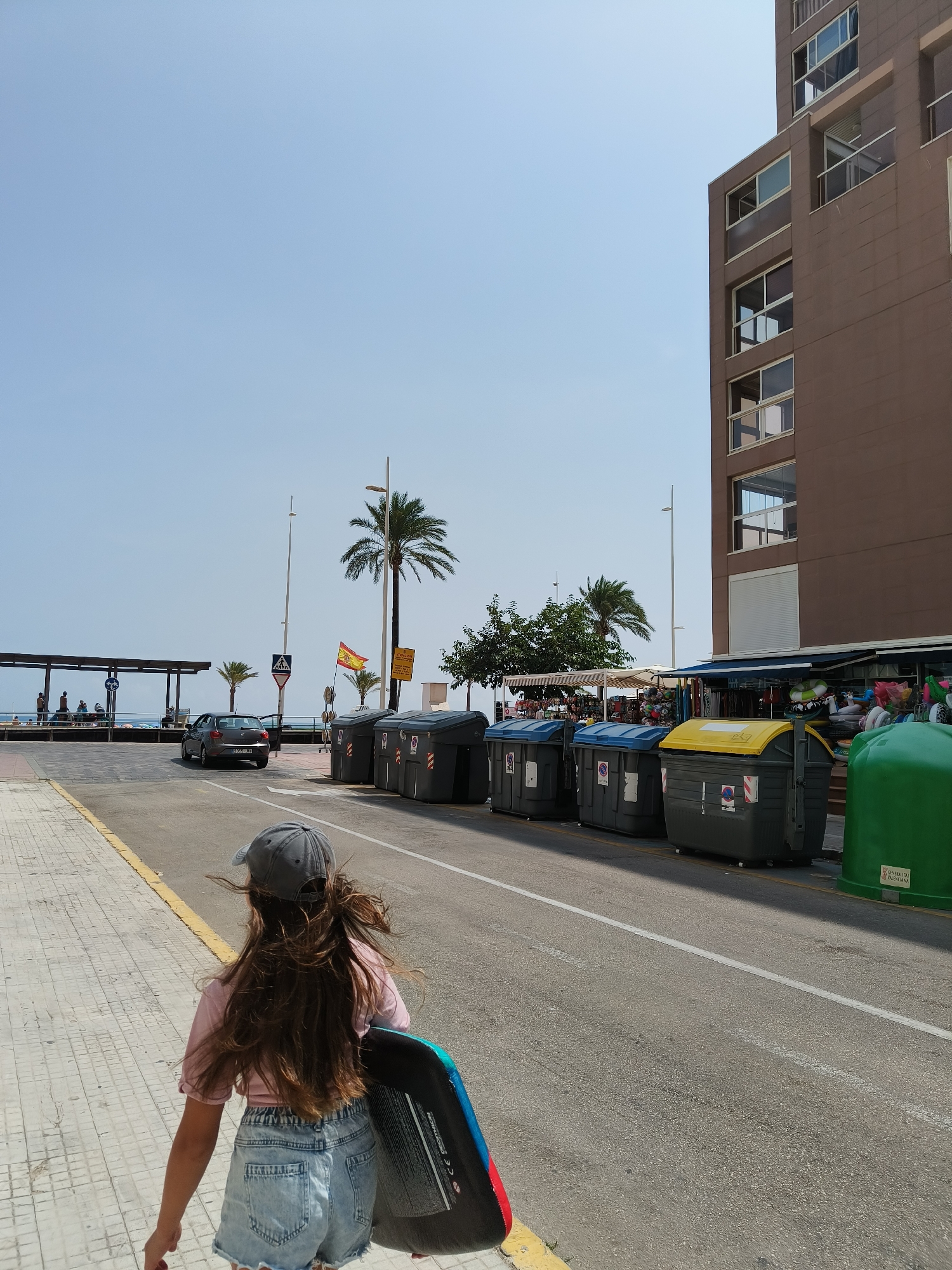 The image shows a scene in Grau I Platja, Spain, on a bright, sunny day with a clear blue sky. In the foreground, a young girl with long brown hair and wearing a grey baseball cap, a pink t-shirt, and denim shorts is walking away from the camera on a tiled sidewalk. She is carrying a blue and black boogie board. To her right, a street runs alongside the sidewalk, where several large, dark grey dumpsters with various colored lids (blue, yellow) are lined up. Further down the street, a grey car is driving away.
On the other side of the street, there is a pedestrian walkway and a covered area with people sitting underneath. Beyond that, the sea is visible. Palm trees are scattered throughout the scene, and a Spanish flag is flying on a pole. To the right of the dumpsters, a tall, brown building with multiple floors and windows is visible. In front of the building, there are shops with awnings and displays of merchandise, including inflatable rings and other beach items. A large green dumpster is positioned to the far right of the image. The overall environment suggests a coastal town or resort area.