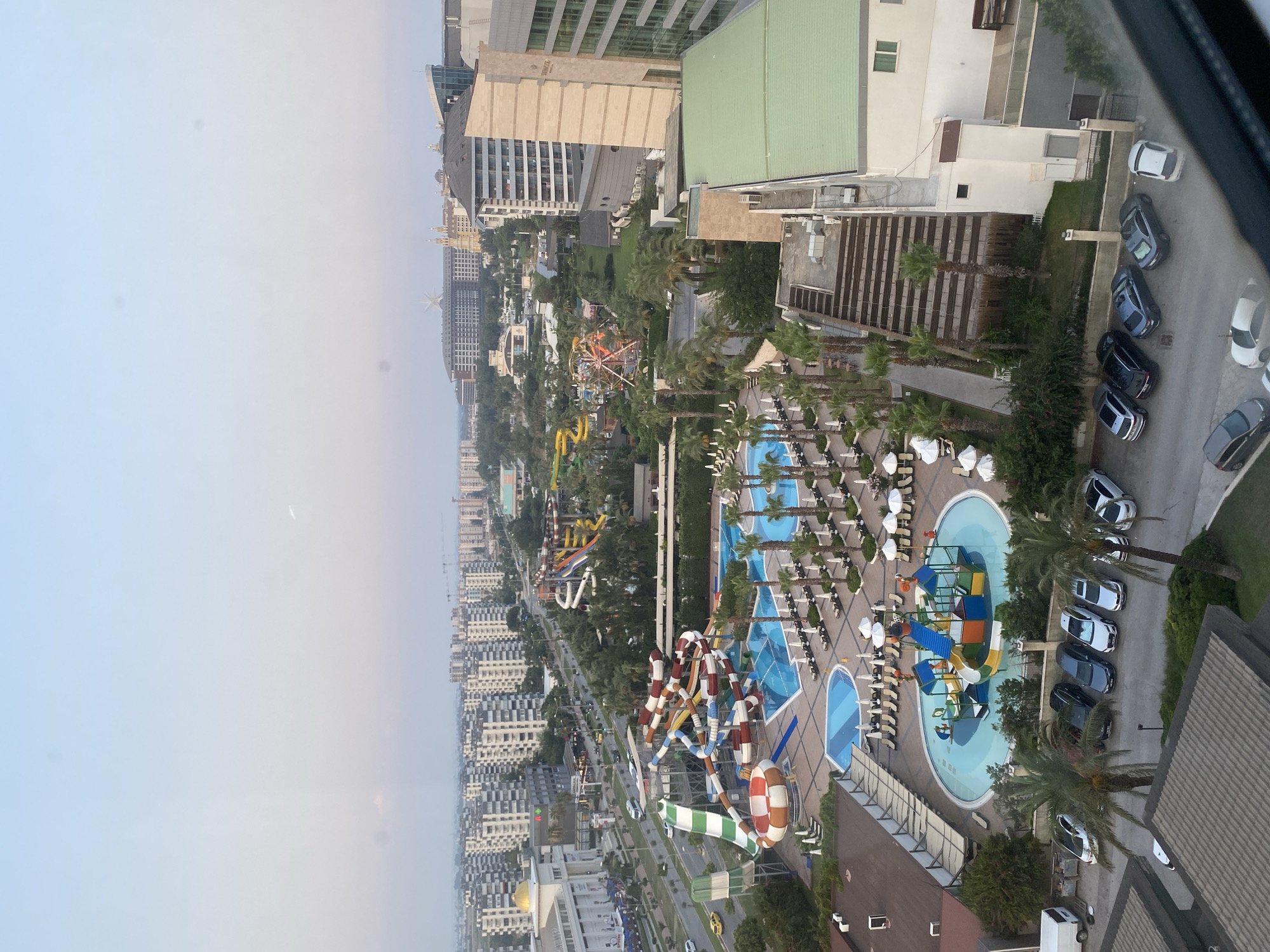 This is a high-angle, medium shot of a resort complex in Aksu, Turkey, likely taken from a hotel room window. The scene depicts a water park with multiple slides, pools, and lounge chairs. Palm trees are interspersed throughout the resort area. Adjacent to the resort are numerous multi-story apartment buildings, typical of a dense urban environment. A road runs alongside the resort, with several cars visible on it and in a parking lot to the left. In the background, more buildings and structures suggest a developed city. The sky is a pale blue with some wispy clouds, indicating it is likely daytime, possibly early morning or late afternoon. There are no discernible people in the image.