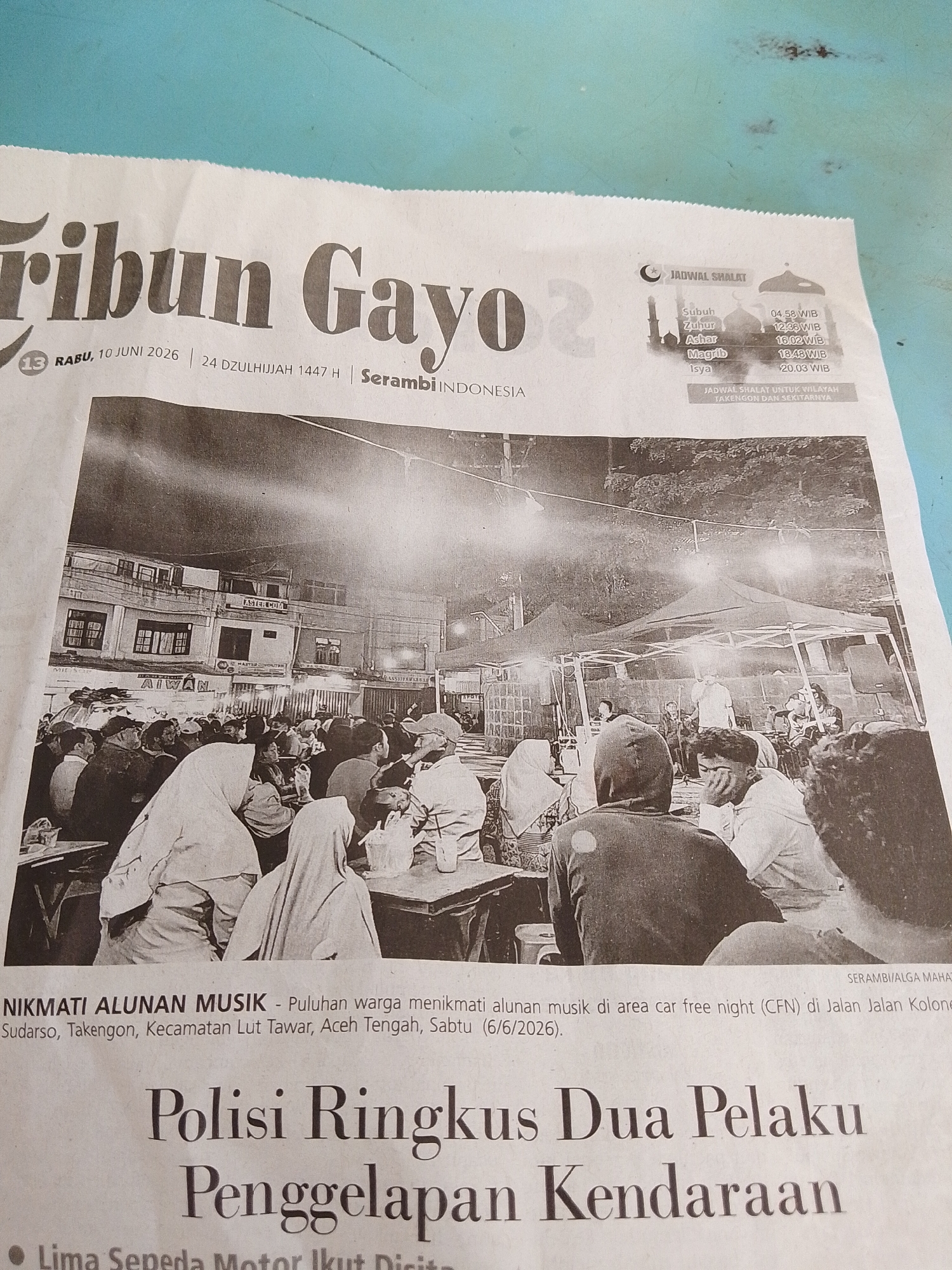The image shows a newspaper article from "Tribun Gayo". The headline reads "Polisi Ringkus Dua Pelaku Penggelapan Kendaraan" (Police Arrest Two Vehicle Embezzlement Perpetrators). There is a sub-headline and caption beneath a photograph that mentions "NIKMATI ALUNAN MUSIK - Puluhan warga menikmati alunan musik di area car free night (CFN) di Jalan Jalan Kolone Sudarso, Takengon, Kecamatan Lut Tawar, Aceh Tengah, Sabtu (6/6/2026)." This translates to "ENJOY THE MUSIC - Dozens of residents enjoy music in the car free night (CFN) area on Jalan Jalan Kolone Sudarso, Takengon, Lut Tawar District, Central Aceh, Saturday (6/6/2026)." The photograph itself depicts a nighttime scene of an outdoor gathering with people seated and a stage with a band performing under tents and lights. There are buildings in the background. The provided crops show details of the newspaper masthead with the date "Rabu, 10 Juni 2026 | 24 Dzullhijjah 1447 H", prayer times, and more of the photograph. There is no information in the image that places this in the city of Lhokseumawe, Indonesia; the location mentioned is Takengon, Central Aceh.