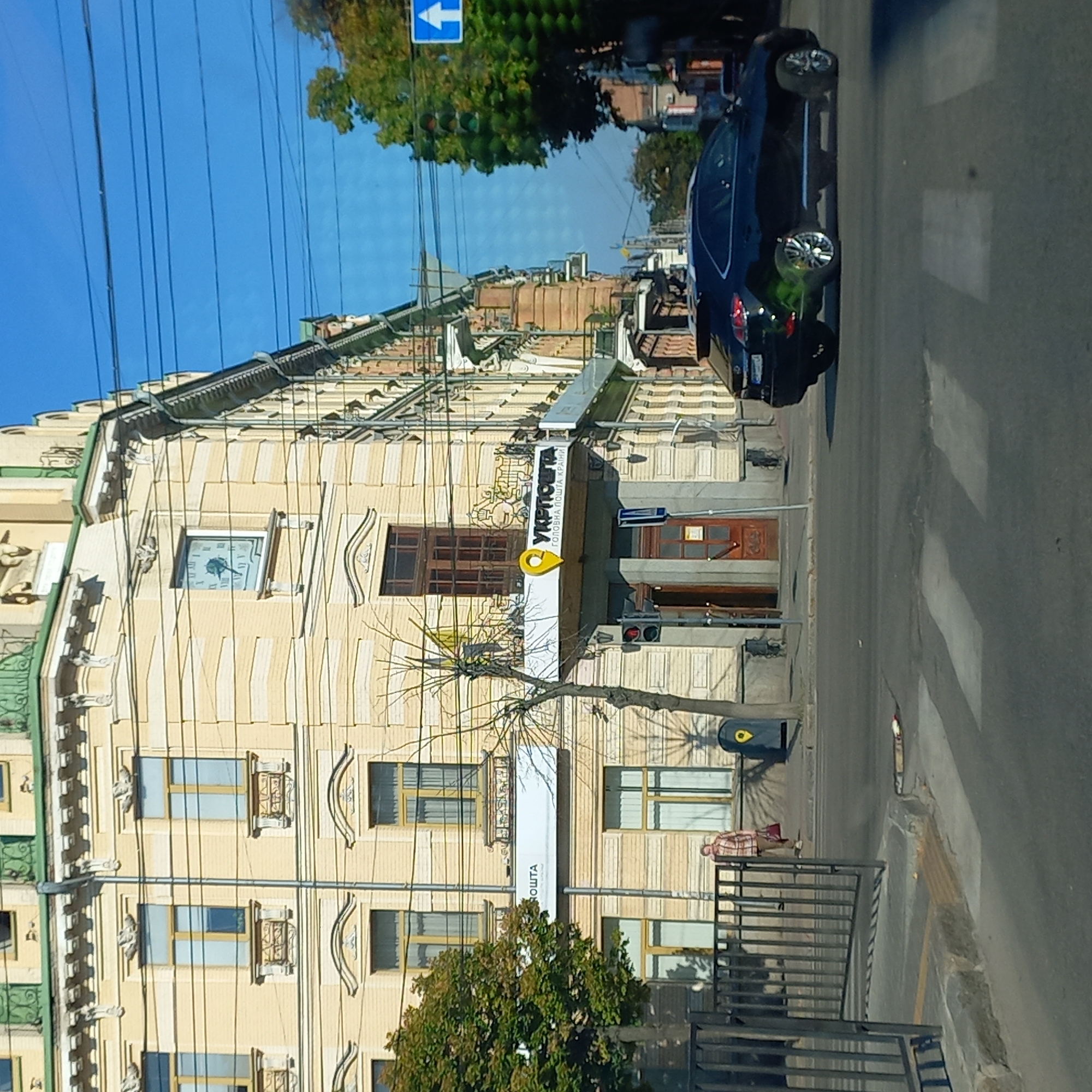 A daytime exterior view captures a segment of a yellow multi-story building with ornate architectural details and visible power lines overhead. A dark-colored SUV is driving across a pedestrian crossing in the foreground. A woman in a plaid shirt and carrying a red bag is standing on the sidewalk near a fence. A sign with a yellow pin icon and Ukrainian text reading "УКРПОШТА" (Ukrposhta) is prominently displayed on the building, indicating a post office. A traffic light is visible on the left side of the frame. The scene is set on a street in Kropyvnytskyi, Ukraine.
